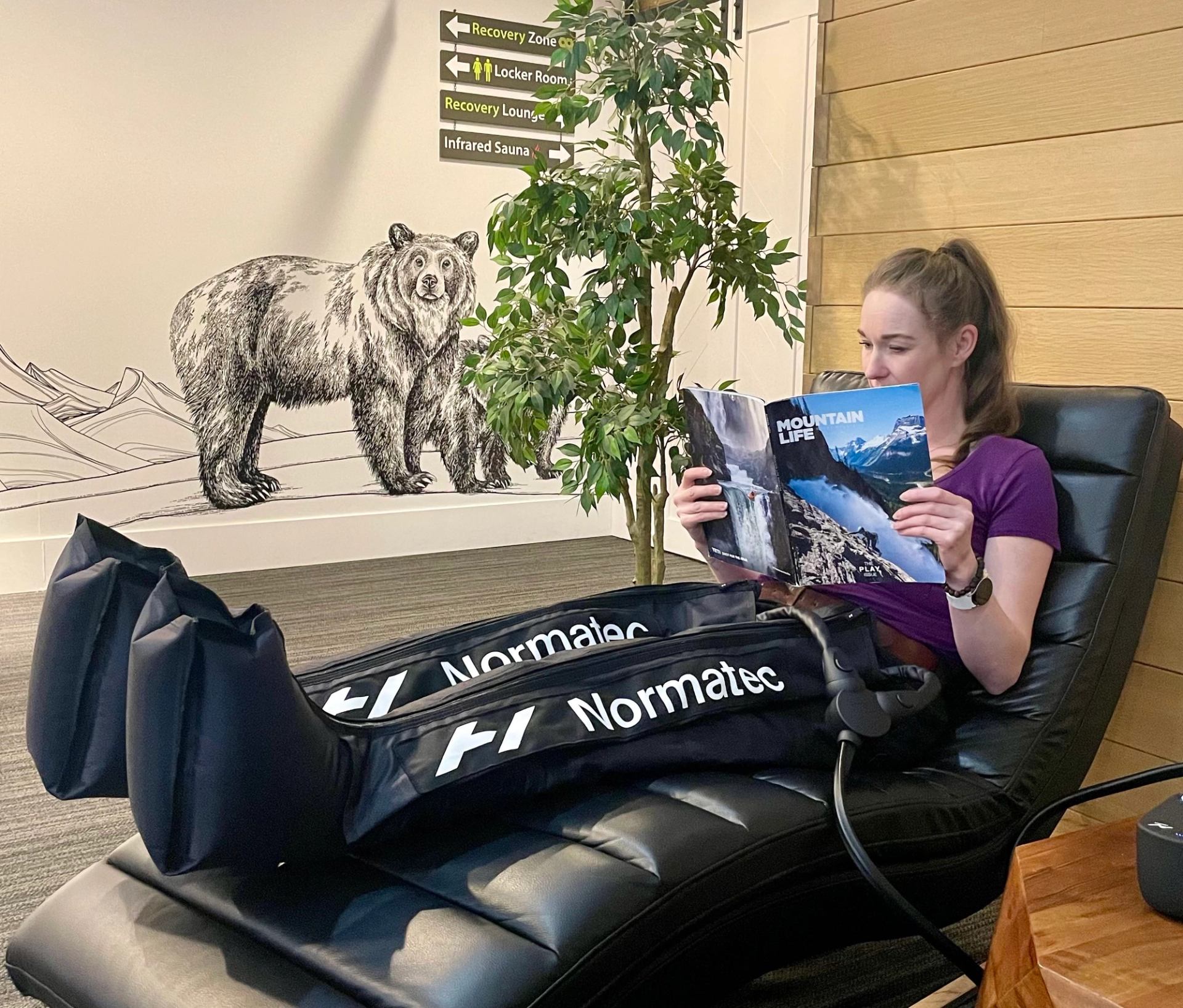 Person relaxing in Normatec boots, reading in a cozy room with a bear mural and plants.