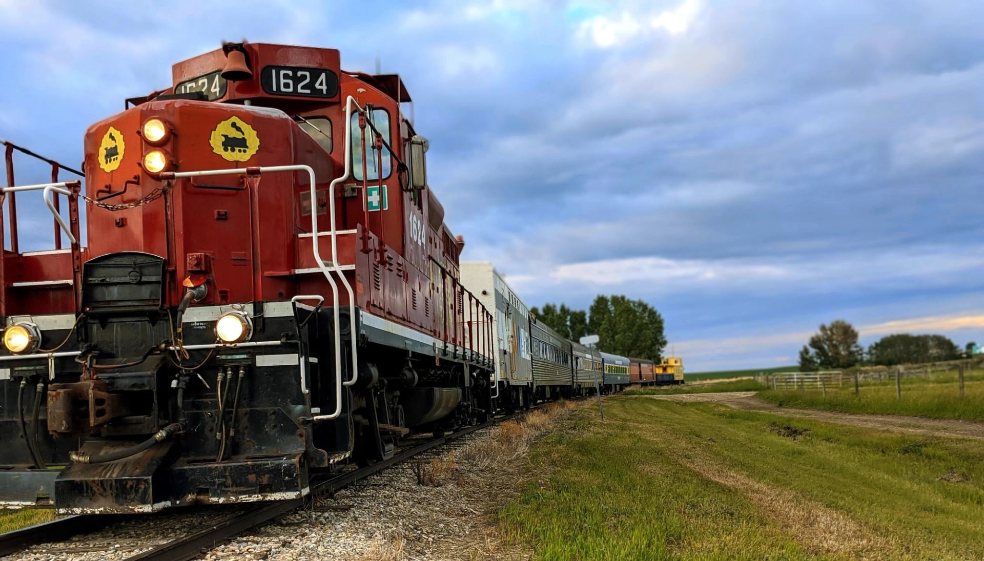 Red locomotive engine 1624 pulls a line of train cars through grassy farmland under cloudy skies.