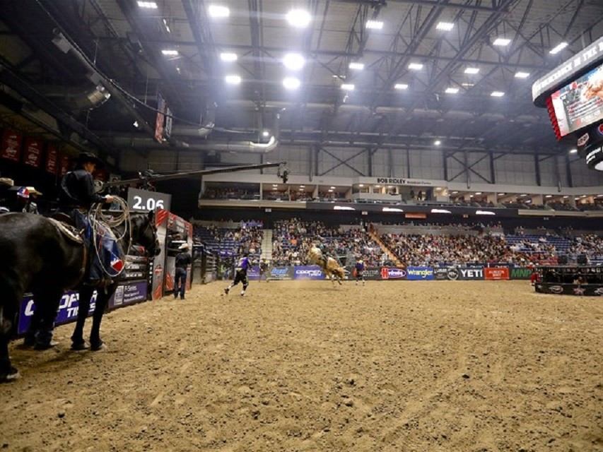 Indoor rodeo arena with dirt floor, spectators, and live horseback action.