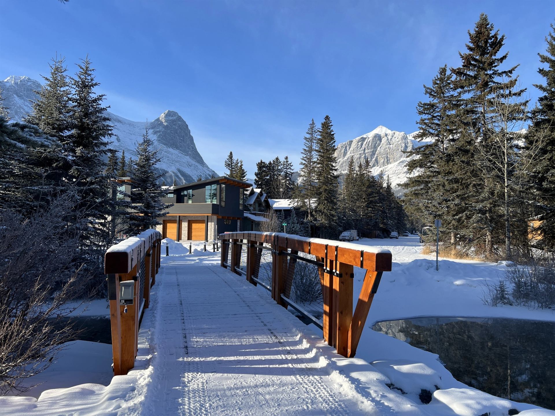 Snow-covered wooden footbridge leading to modern houses, evergreen trees and distant mountains.