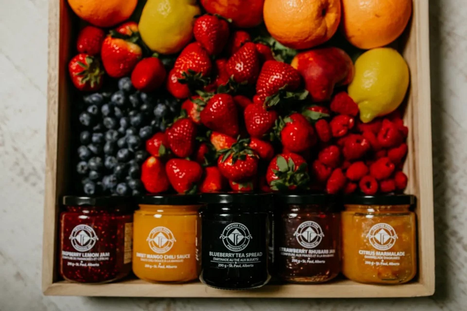 Wooden tray with assorted fresh berries, citrus fruits and five labelled jars of preserves.