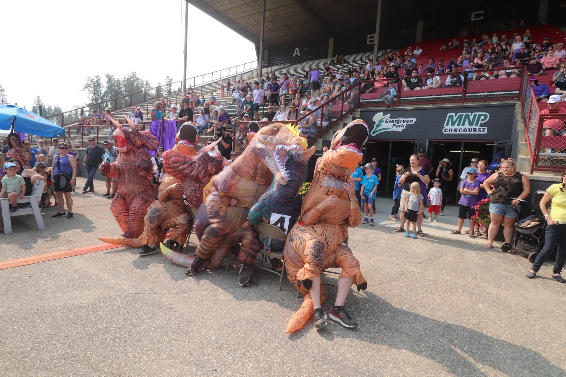 People in inflatable dinosaur costumes sit on chairs as crowd watches at Evergreen Park.