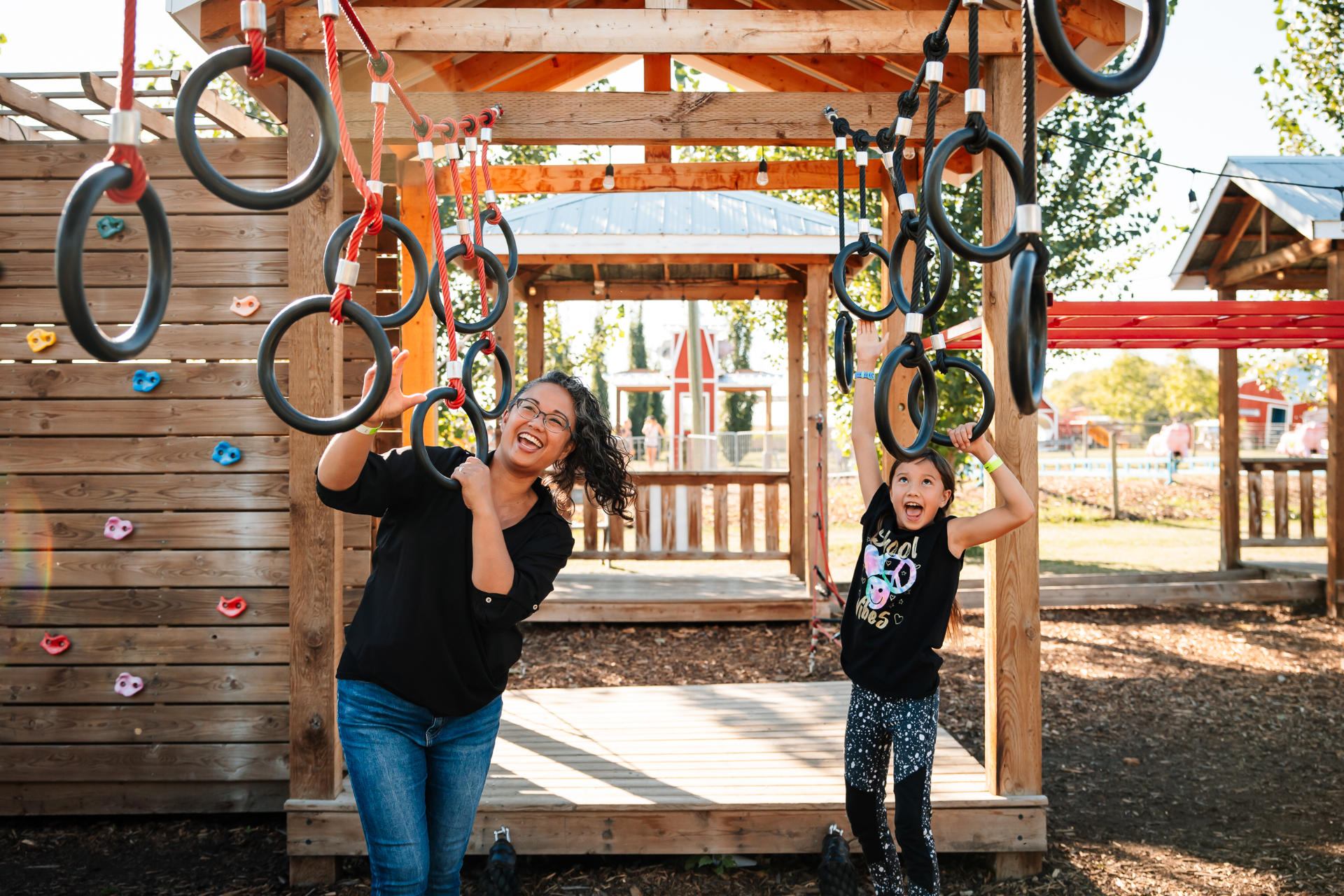Two people hang from playground rings under a wooden structure with climbing wall.