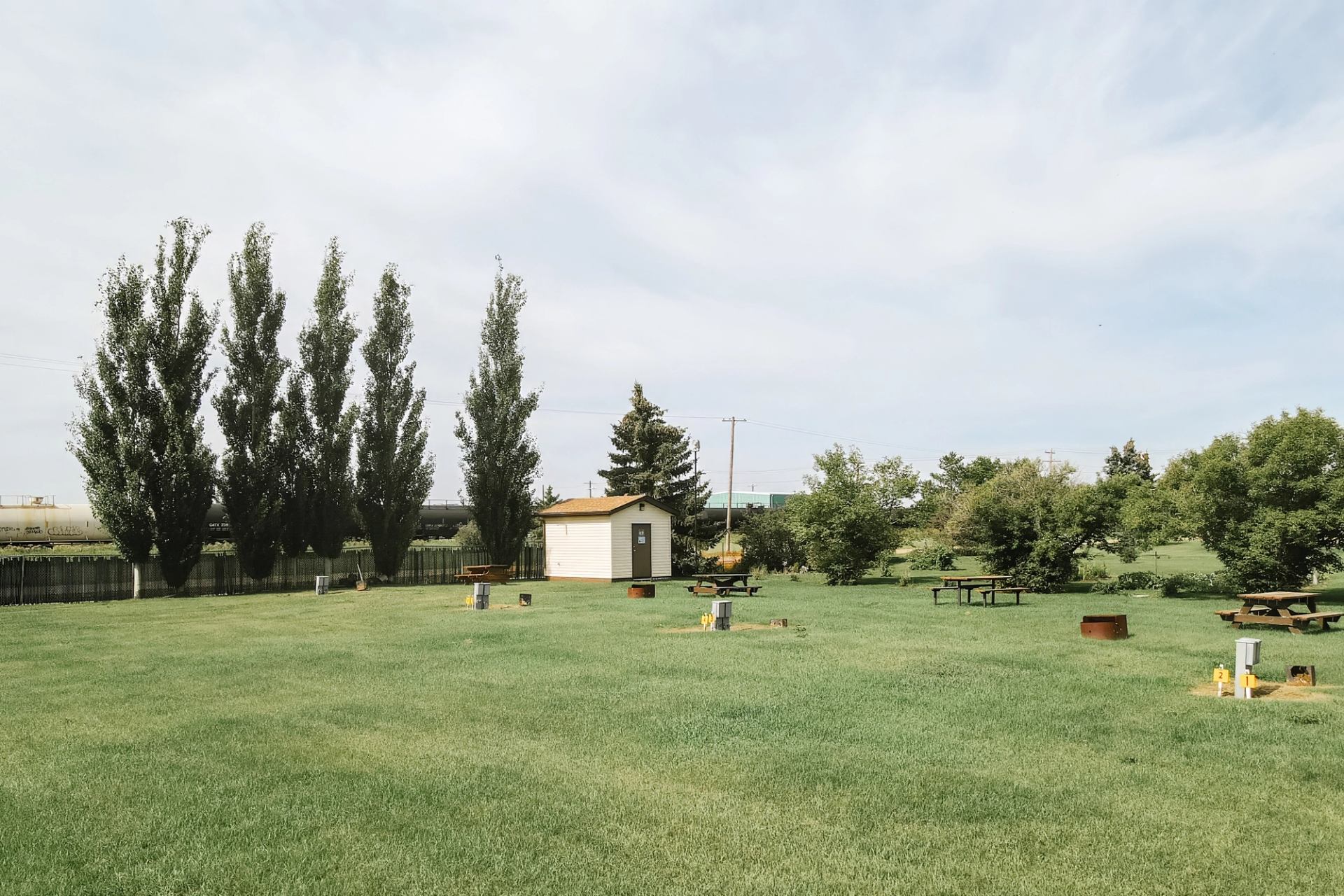 Grassy campground with picnic tables, fire pits and small utility hookups near trees.