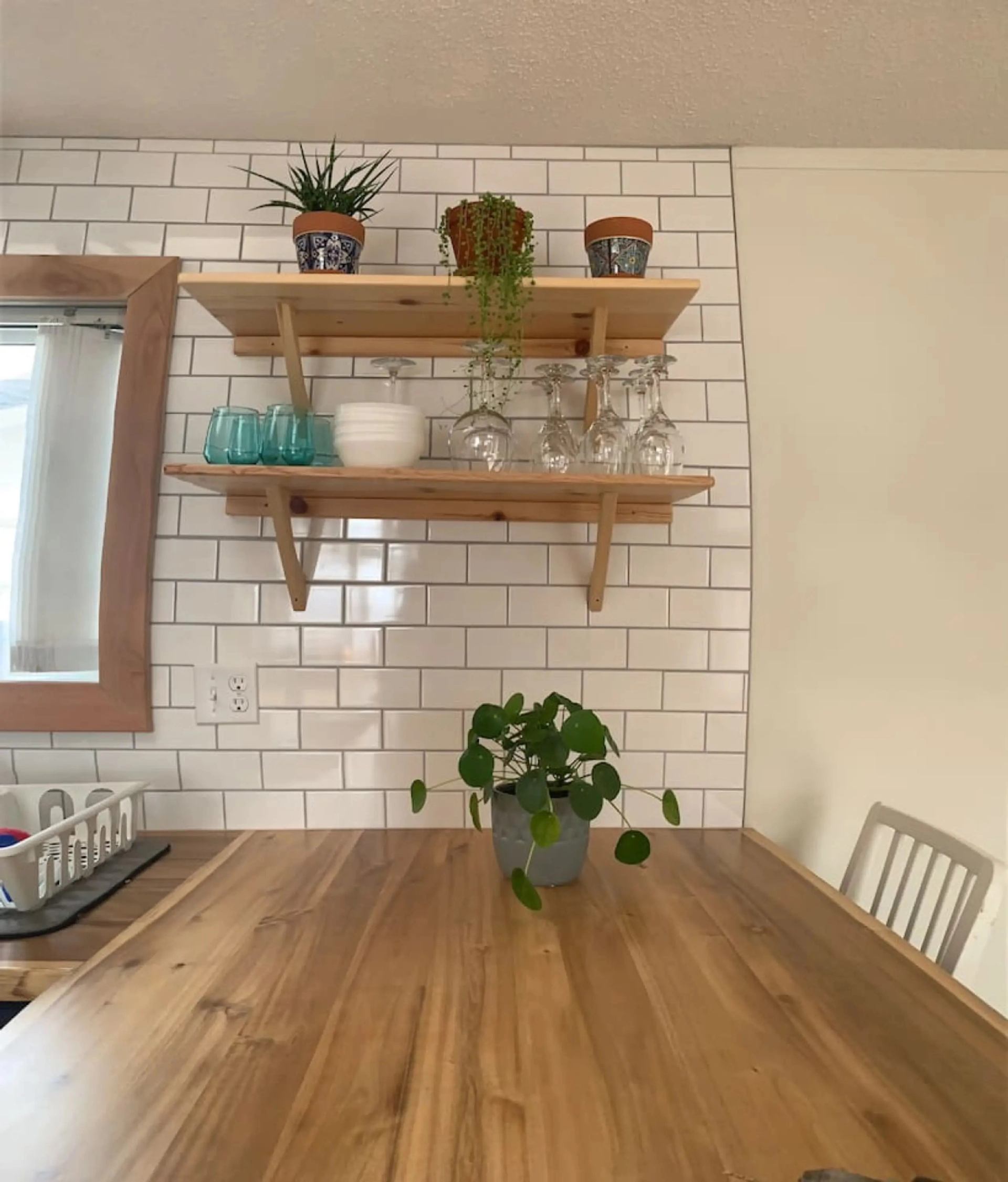 Wood dining table with potted plant beneath shelves of glassware and plants.