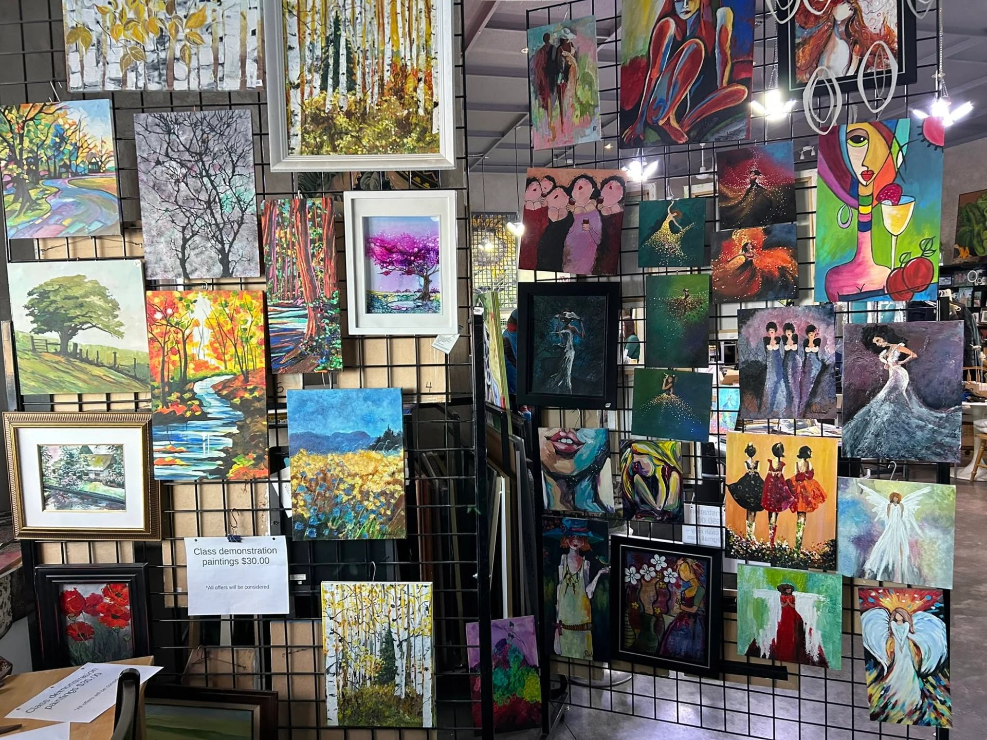 Colourful landscape and figure paintings hang on black grid panels at an art display.