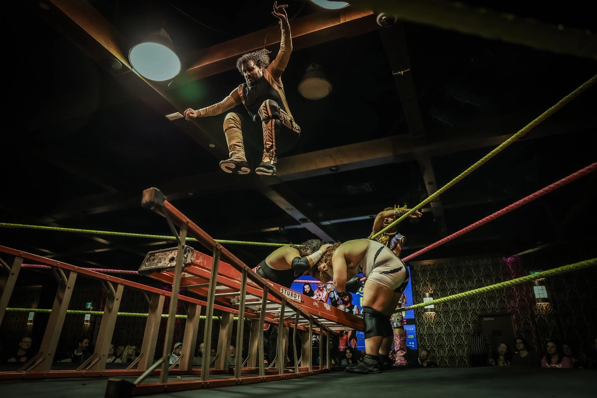 Wrestler jumps from above a ladder toward opponents positioned inside the ring.