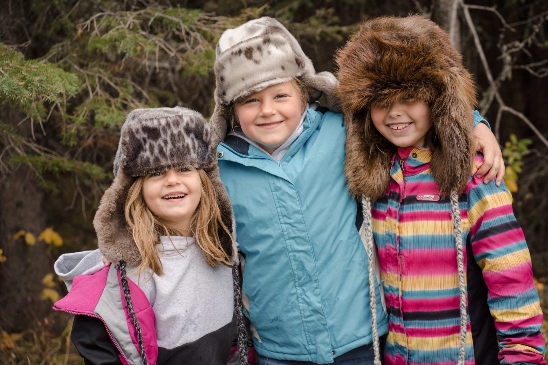 Three smiling children in winter coats and fur hats pose arm-in-arm in a forested outdoor setting.