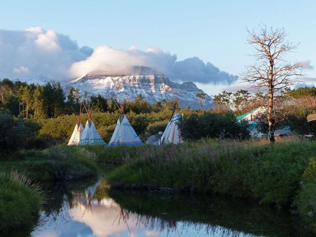 Teepees sit along the edge of a stream with mountains in the background.
