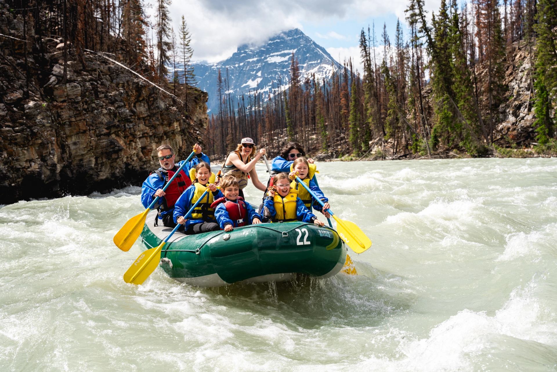 Group rafting through fast river rapids with mountains and forest in the background.
