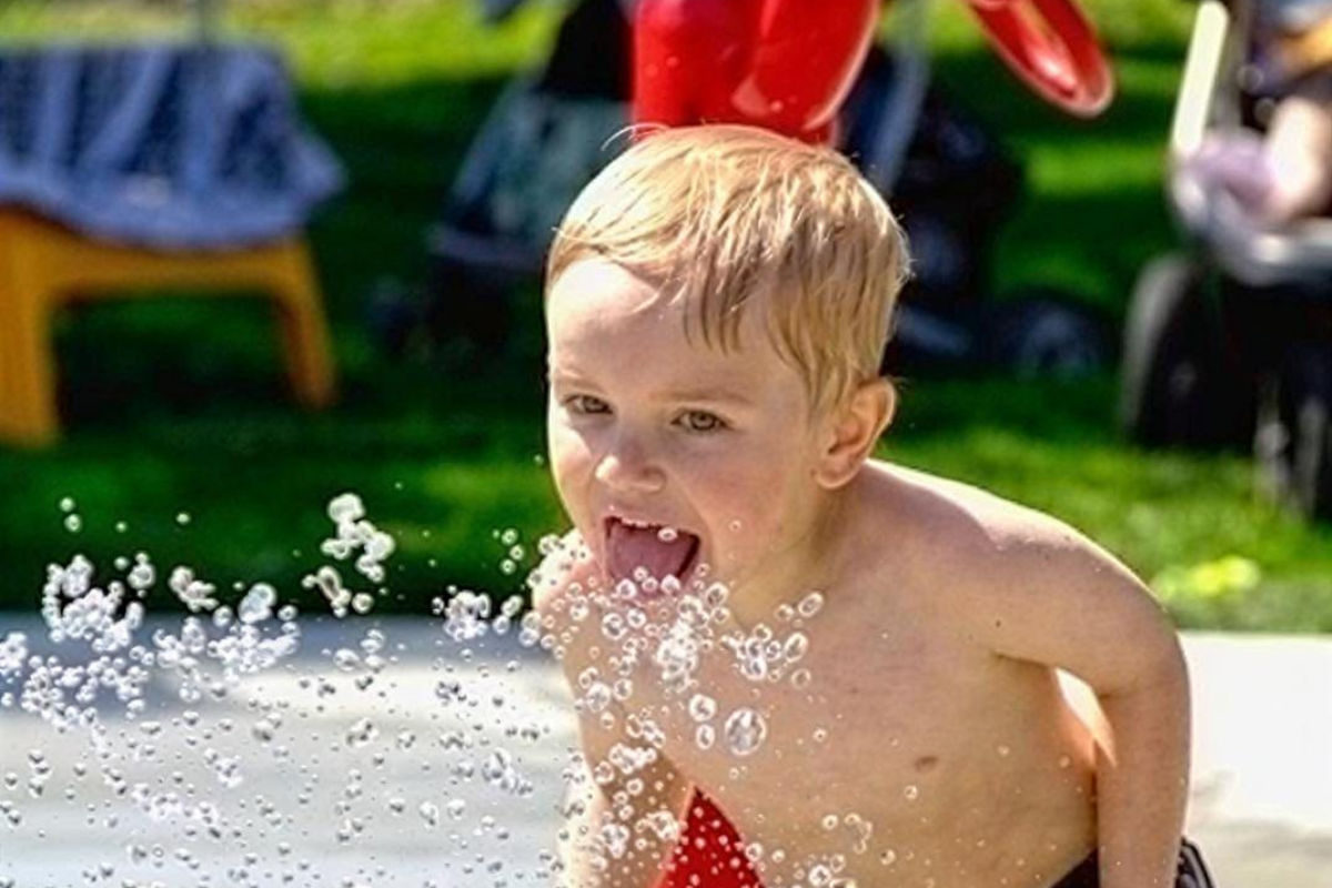 Diamond Valley Municipal Campground showcases a close-up of a child playing in a water feature, with clear droplets splashing in mid-air. Bright outdoor seating and green lawn areas in the background highlight a family-friendly, sunny recreational space.
