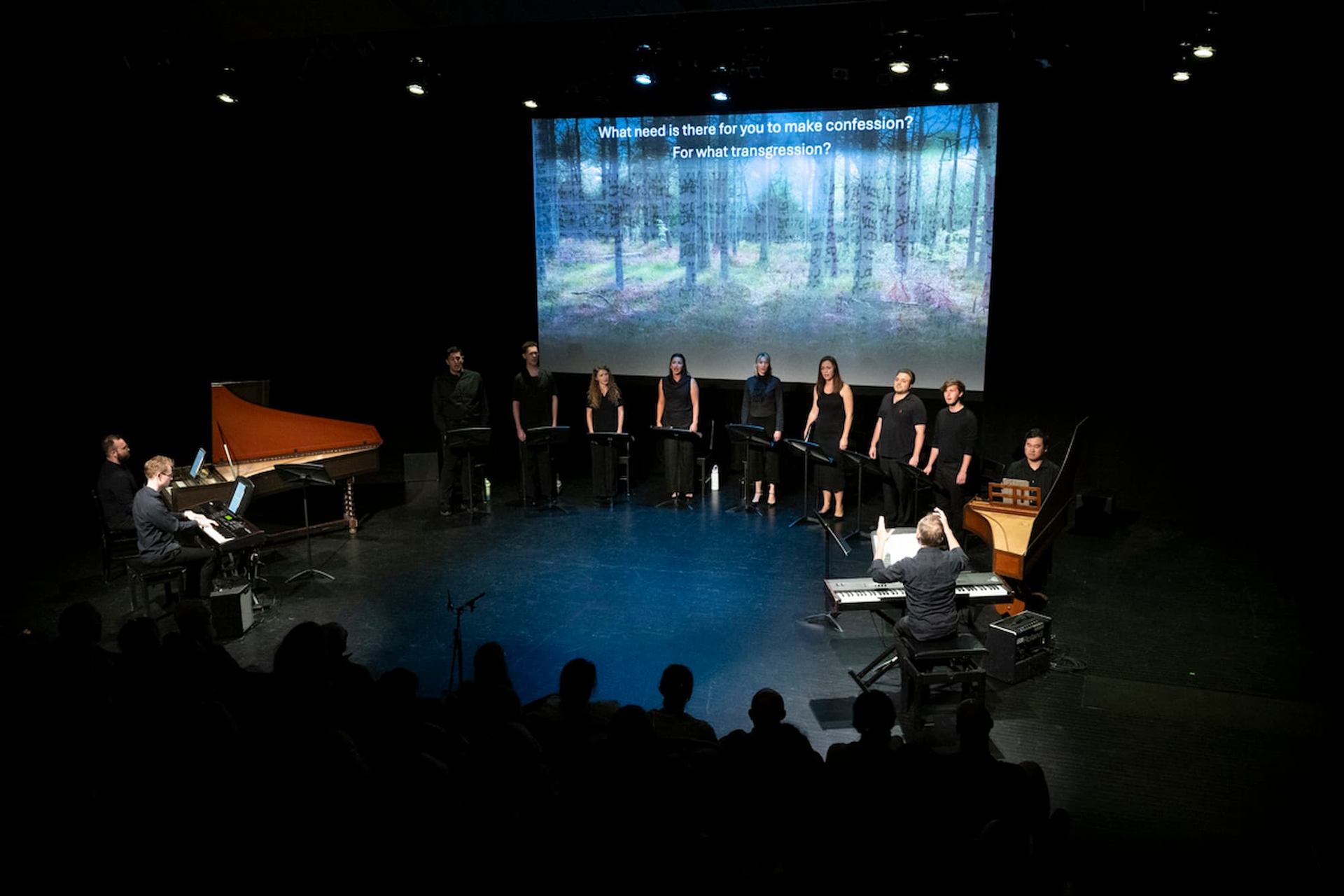 Choir and musicians perform on dark stage before forest projection with superimposed text.