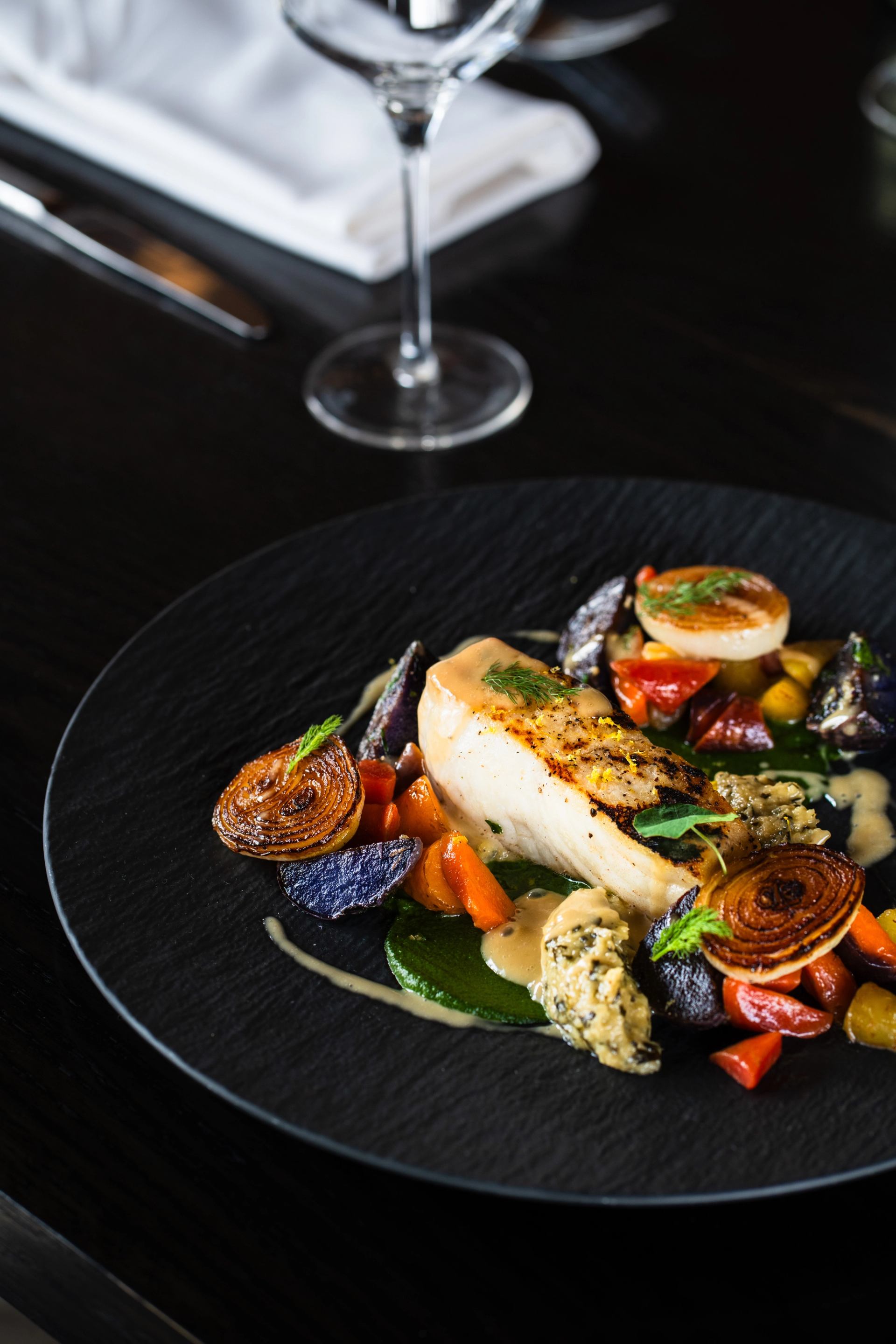 Plated fish fillet with roasted vegetables and sauce on a dark restaurant table.