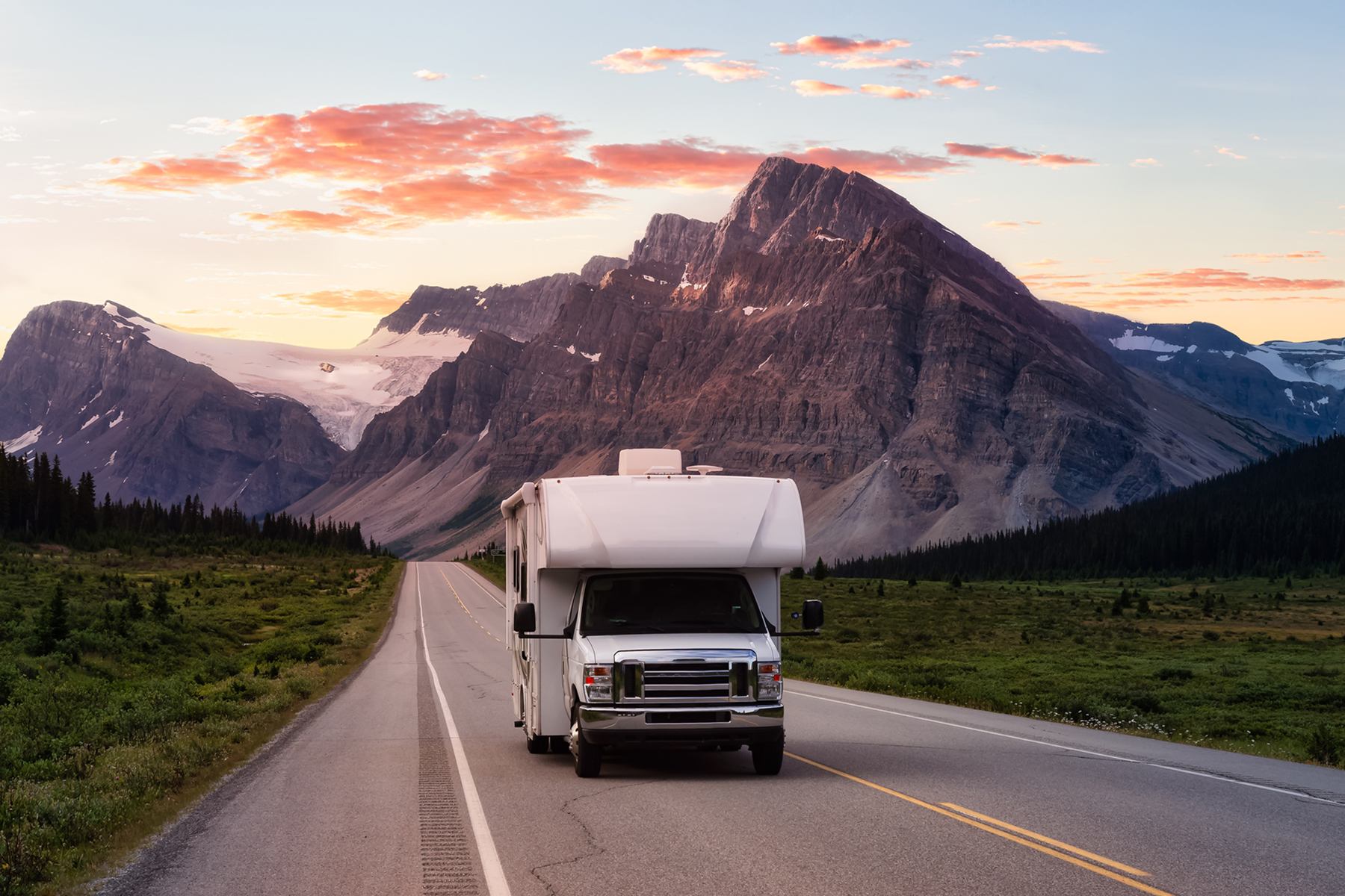 RV driving toward mountains at sunset on a scenic road lined with greenery.