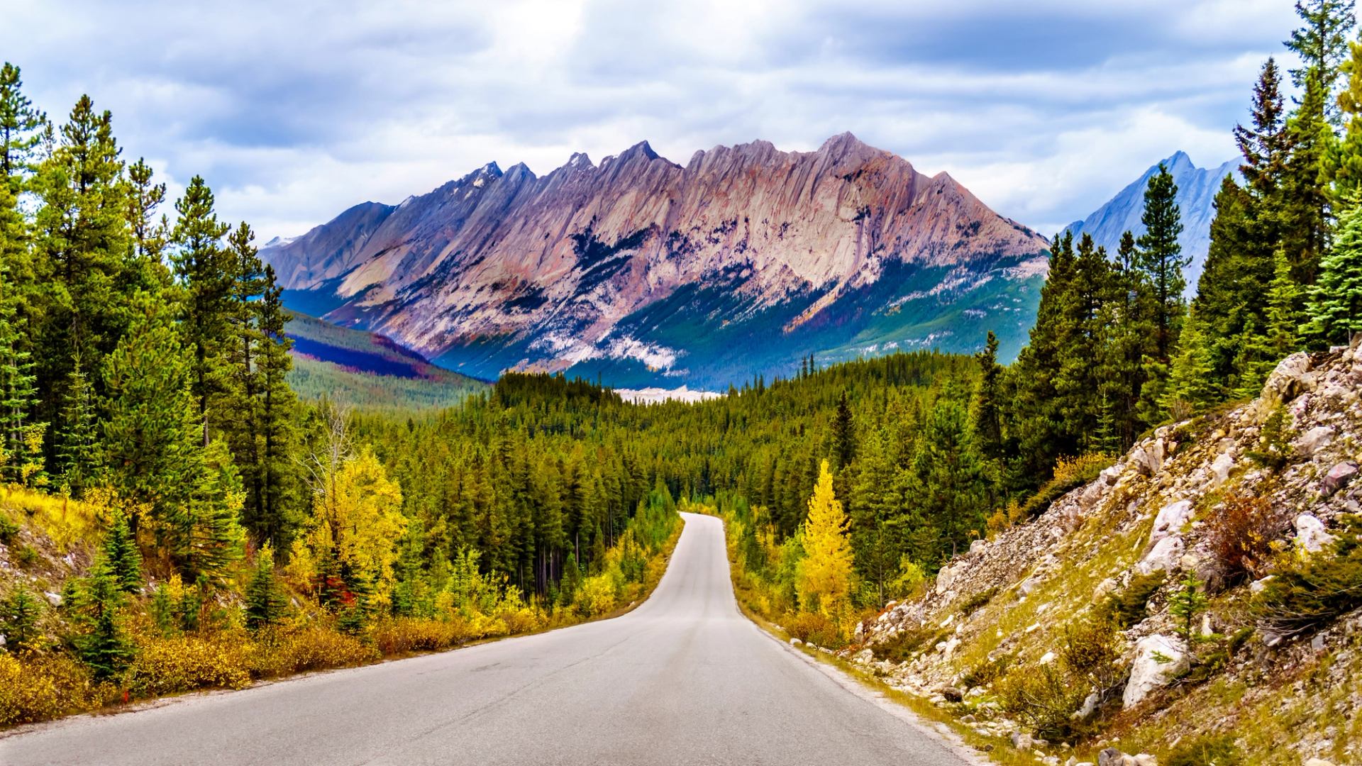 Mountain road surrounded by lush green forest and rugged peaks under a partly cloudy sky.