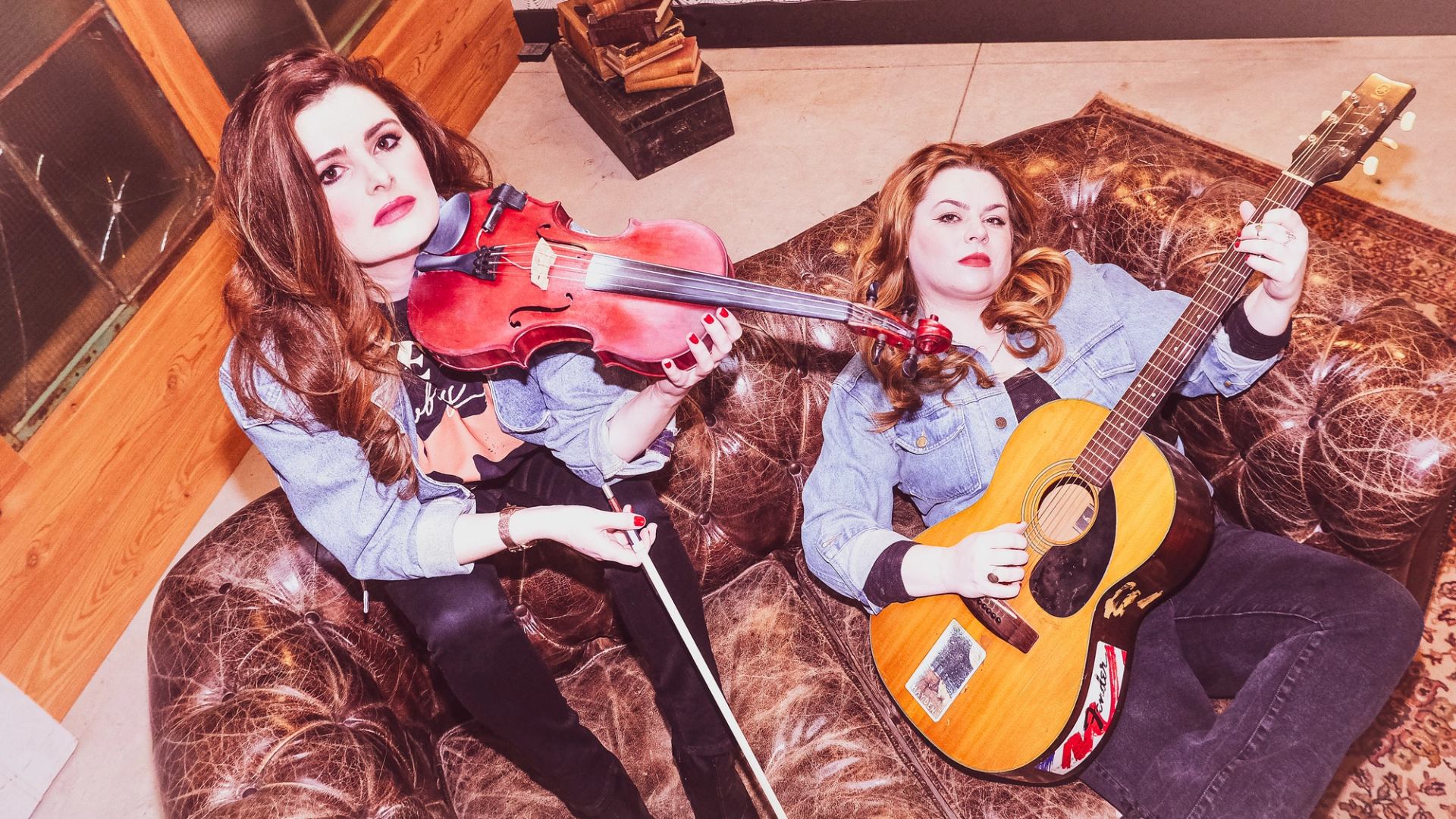 Two people in denim jackets play violin and acoustic guitar on a worn leather couch.