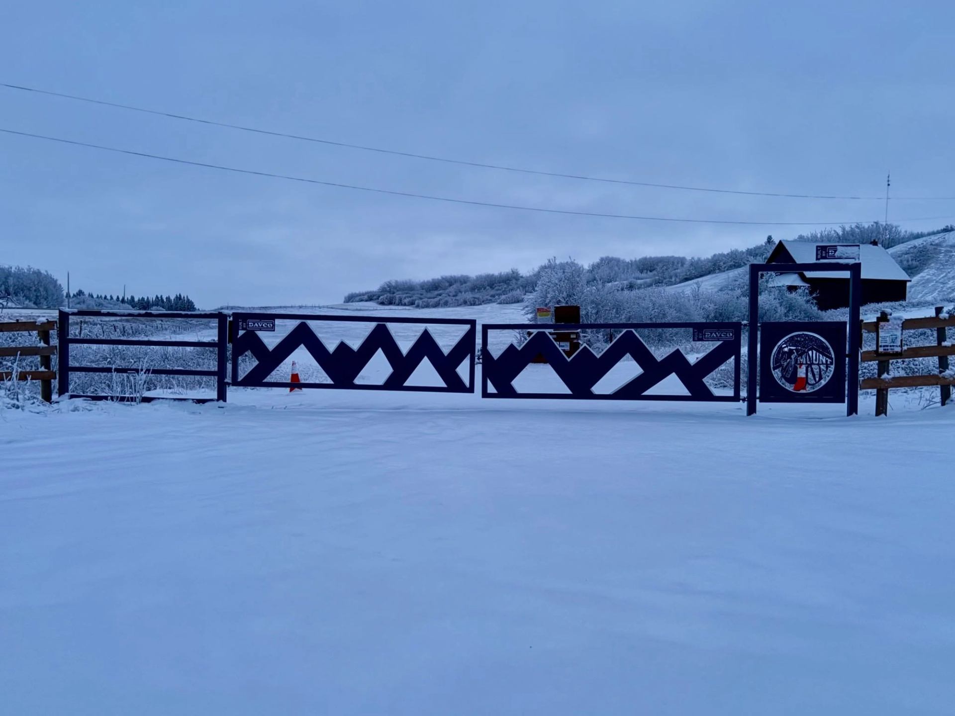 Snow-covered metal gate with zigzag pattern blocking rural road and frosty hills.