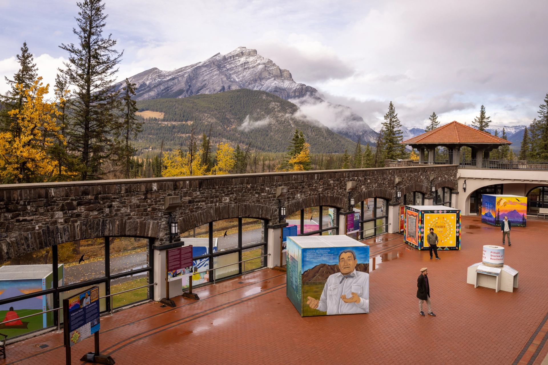 Outdoor courtyard with colourful art installations, people walking, and mountains in the background.