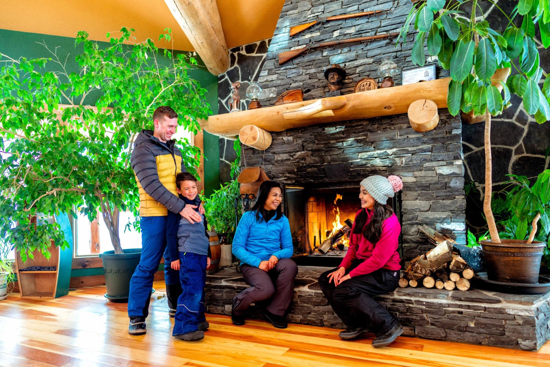 Family relaxing by a large stone fireplace inside a cozy lodge in the Rocky Mountains.