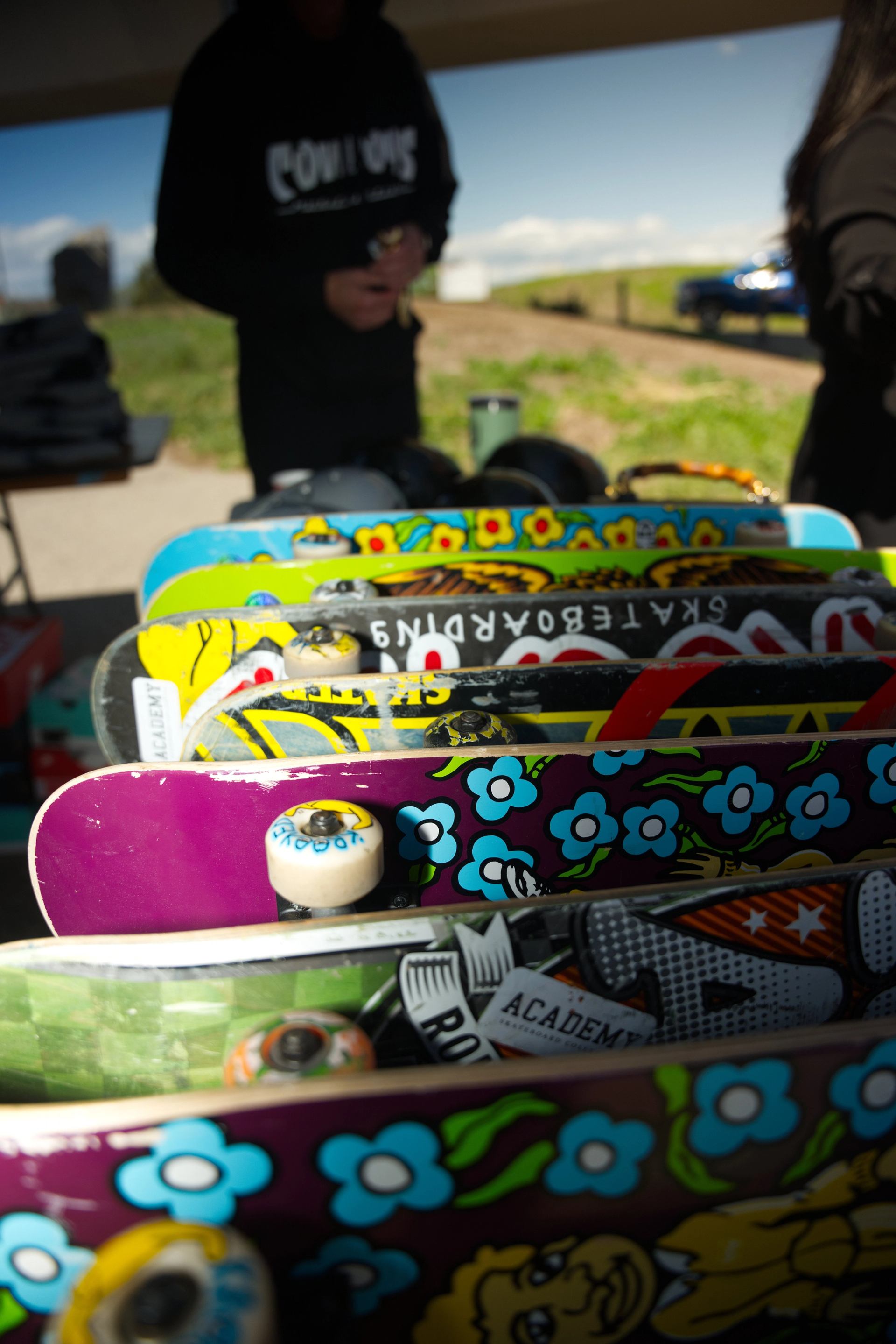 Colourful skateboard decks with cartoon graphics stacked on a table under outdoor shelter.