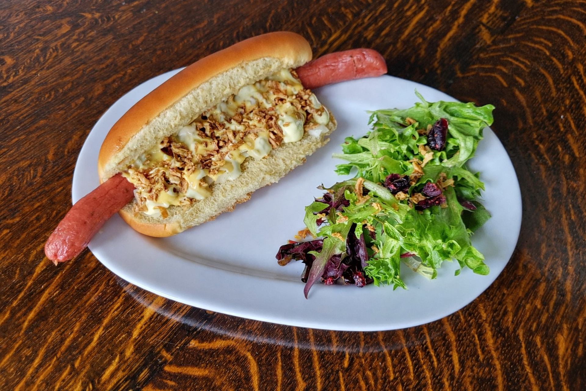 Hot dog with toppings and mixed green salad on white plate over wood table.