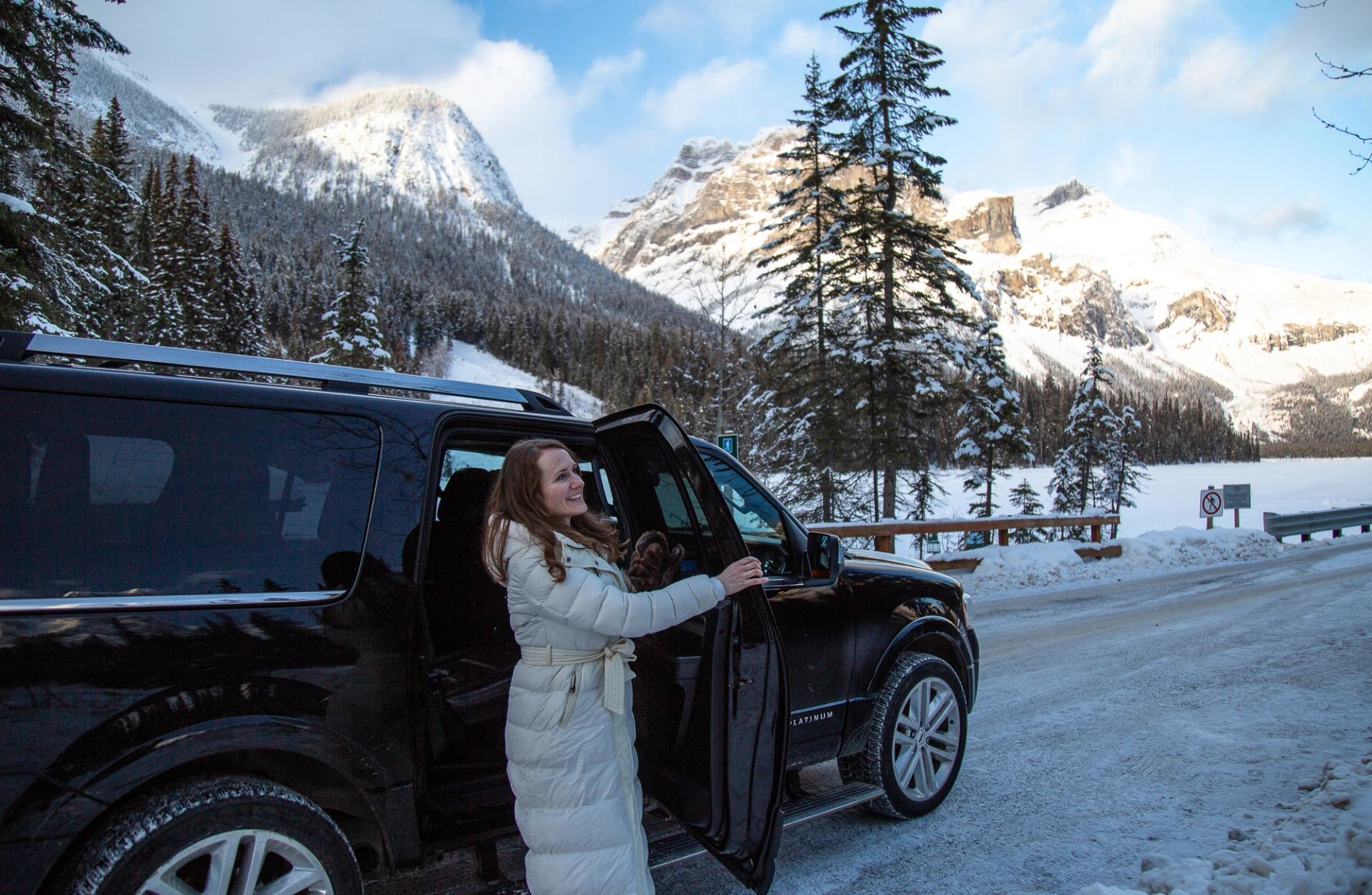 Person standing by a black SUV on a snow-lined road with rugged mountains in the distance
