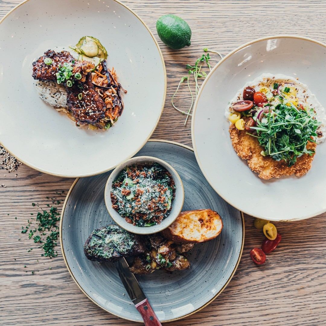 Three plated dishes with meats, greens, and garnishes on a wooden table.