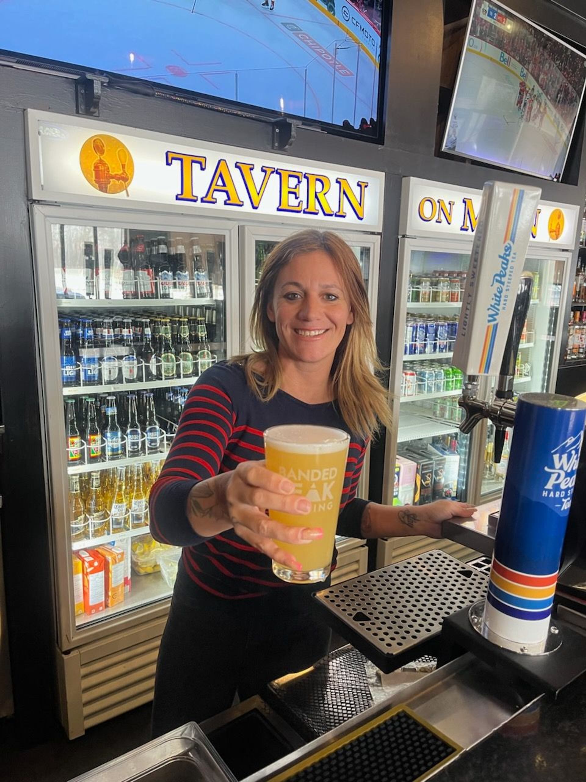 A staff member stands behind a bar, holding out a glass of beer toward the viewer. Behind the counter are glass-door coolers stocked with drinks and signage reading “Tavern on Main,” with TV screens overhead showing sports.