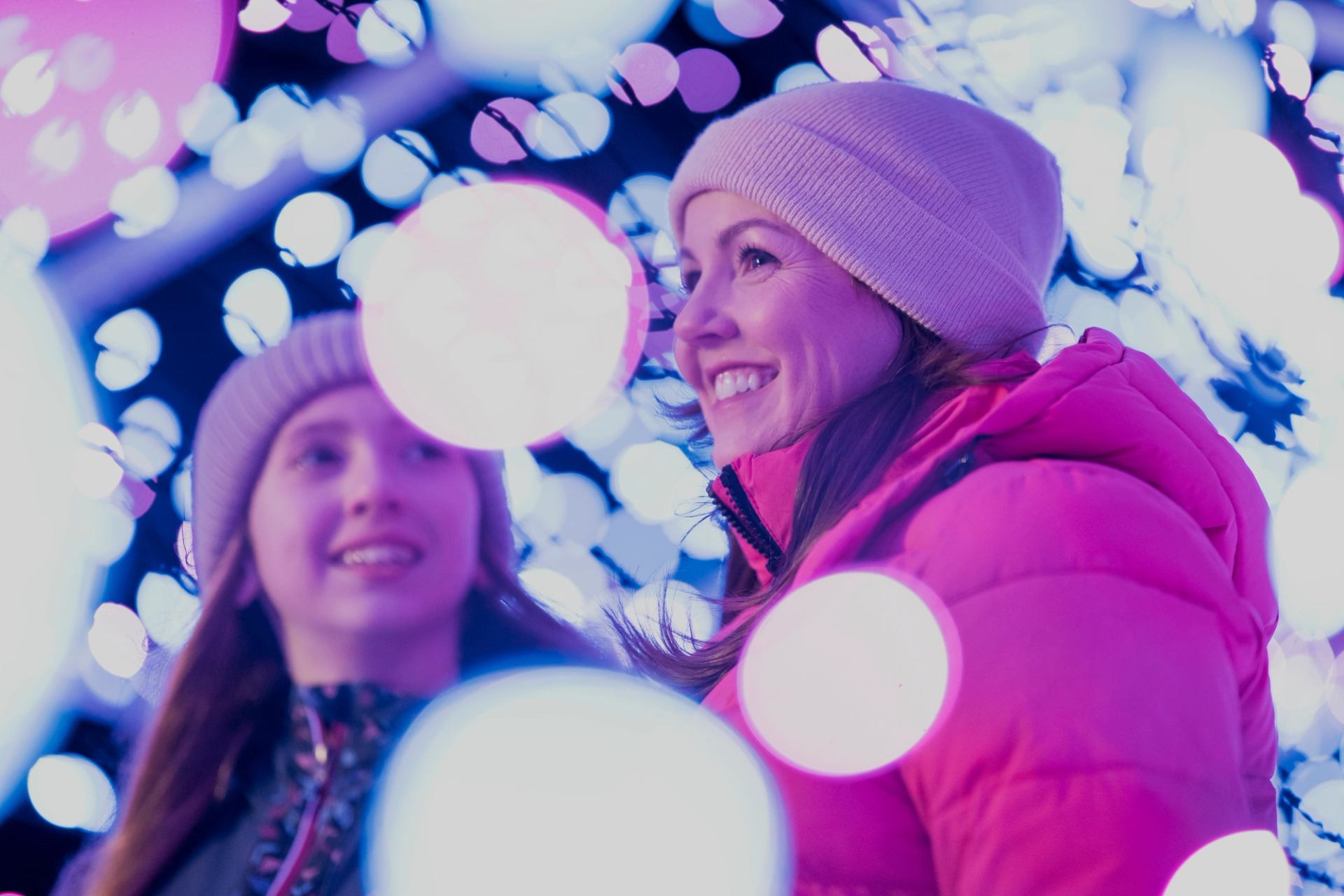 Two people in winter clothing surrounded by bright, colorful festival lights.