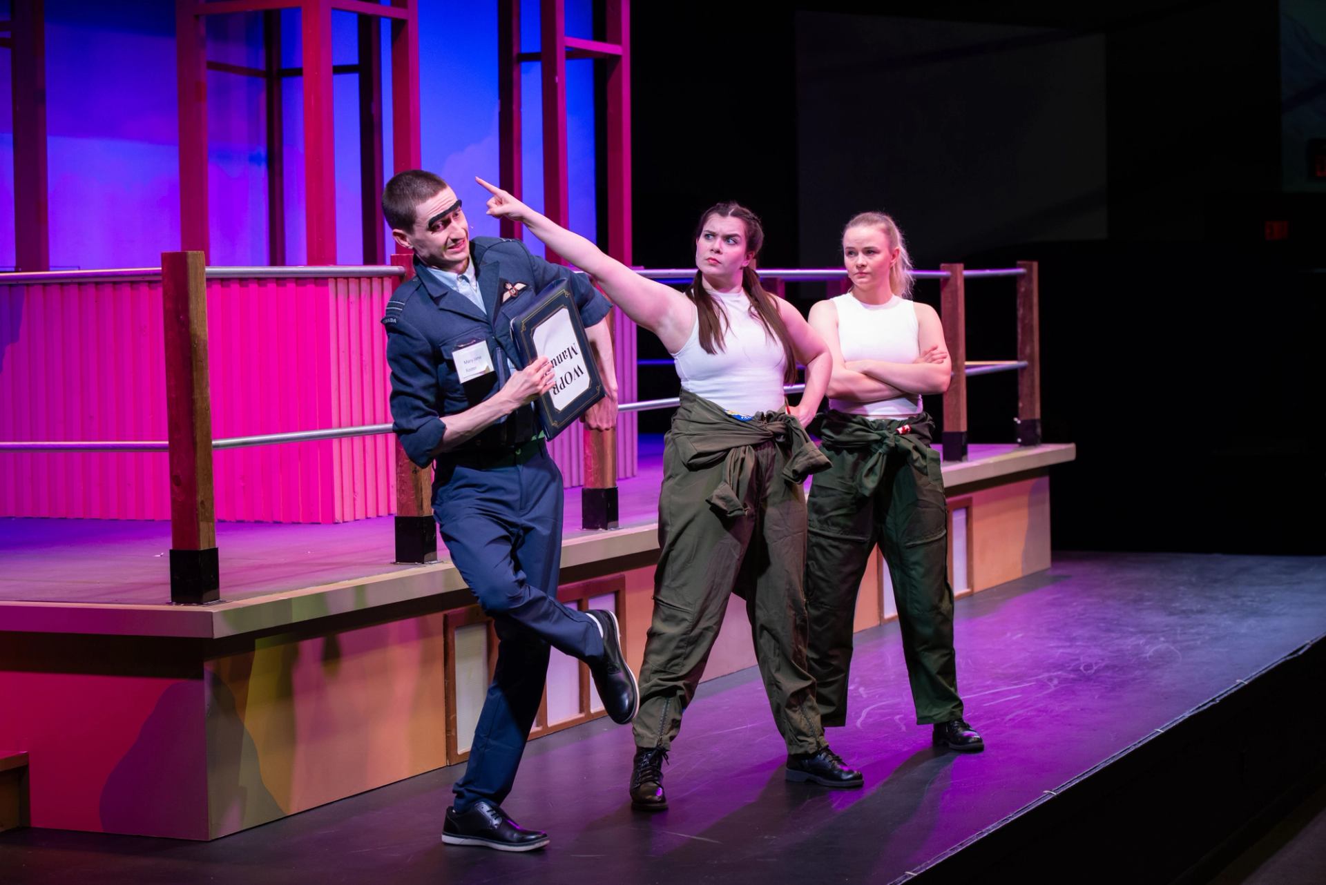 Performer in uniform gesturing while two others in flight suits watch on a colorful stage