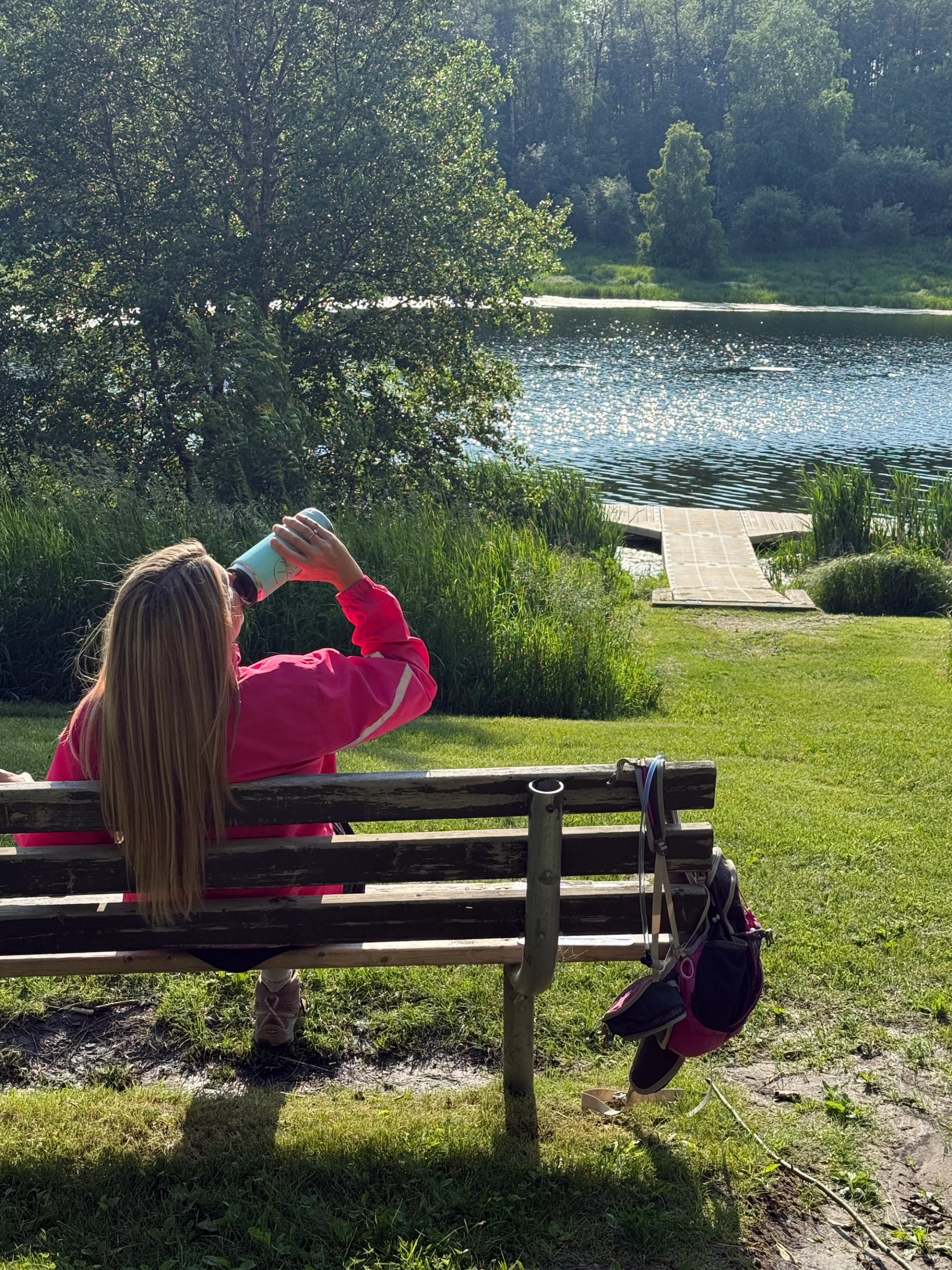 A person sitting on a bench by a lake drinking from a bottle.
