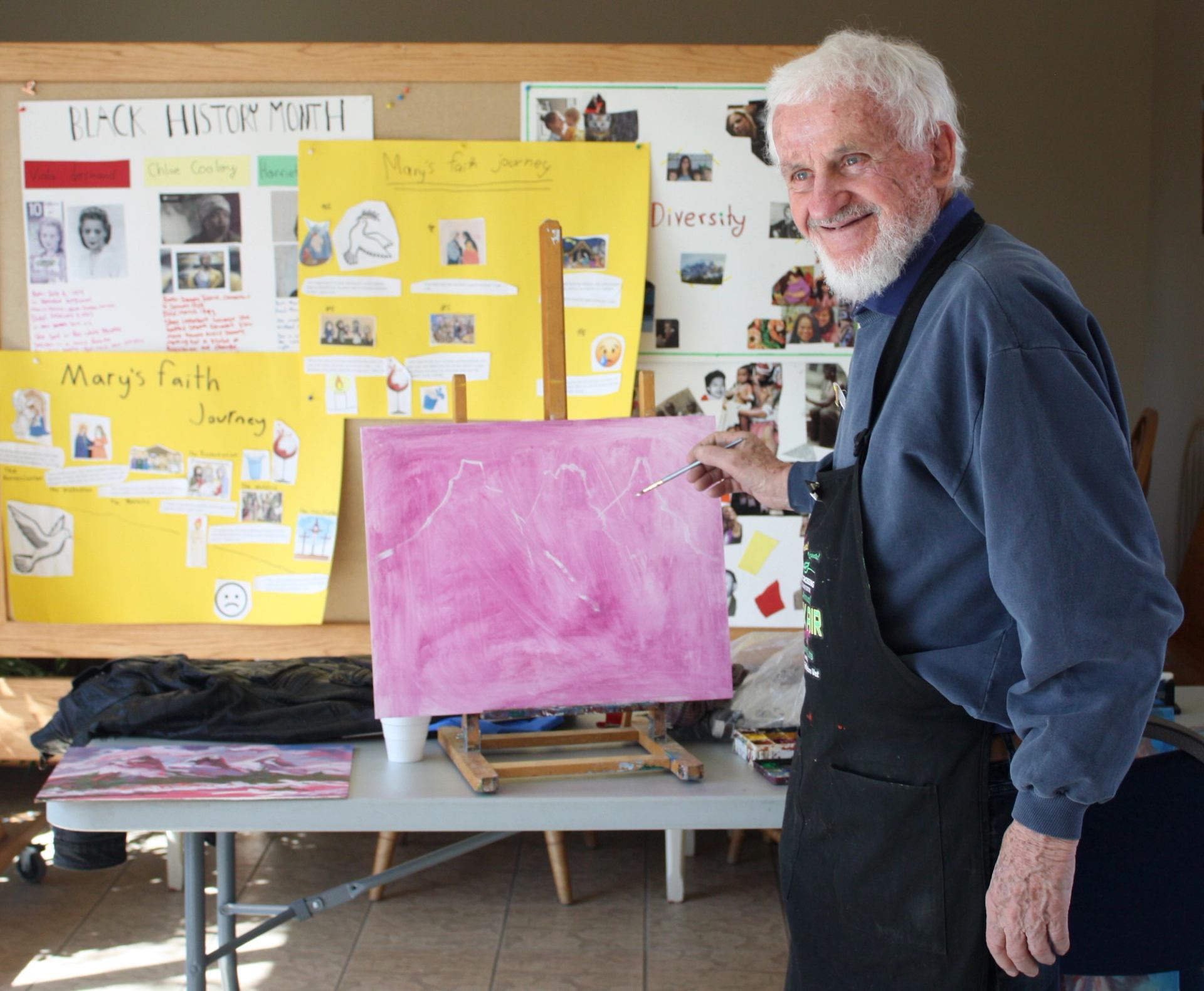 Artist painting pink canvas in classroom with Black History Month and diversity posters behind.