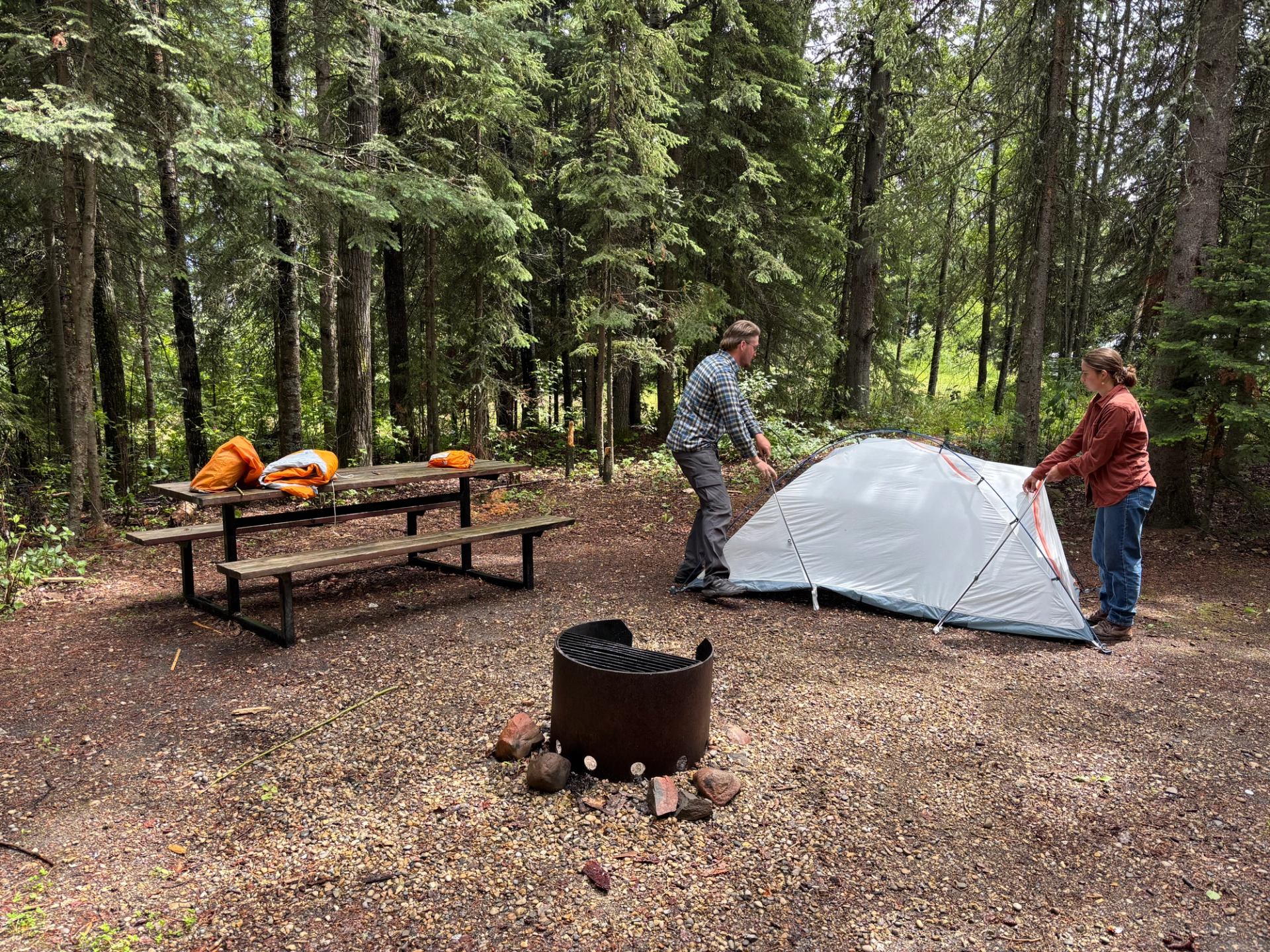 Two people setting up a tent at a forest campsite with a picnic table and fire ring.