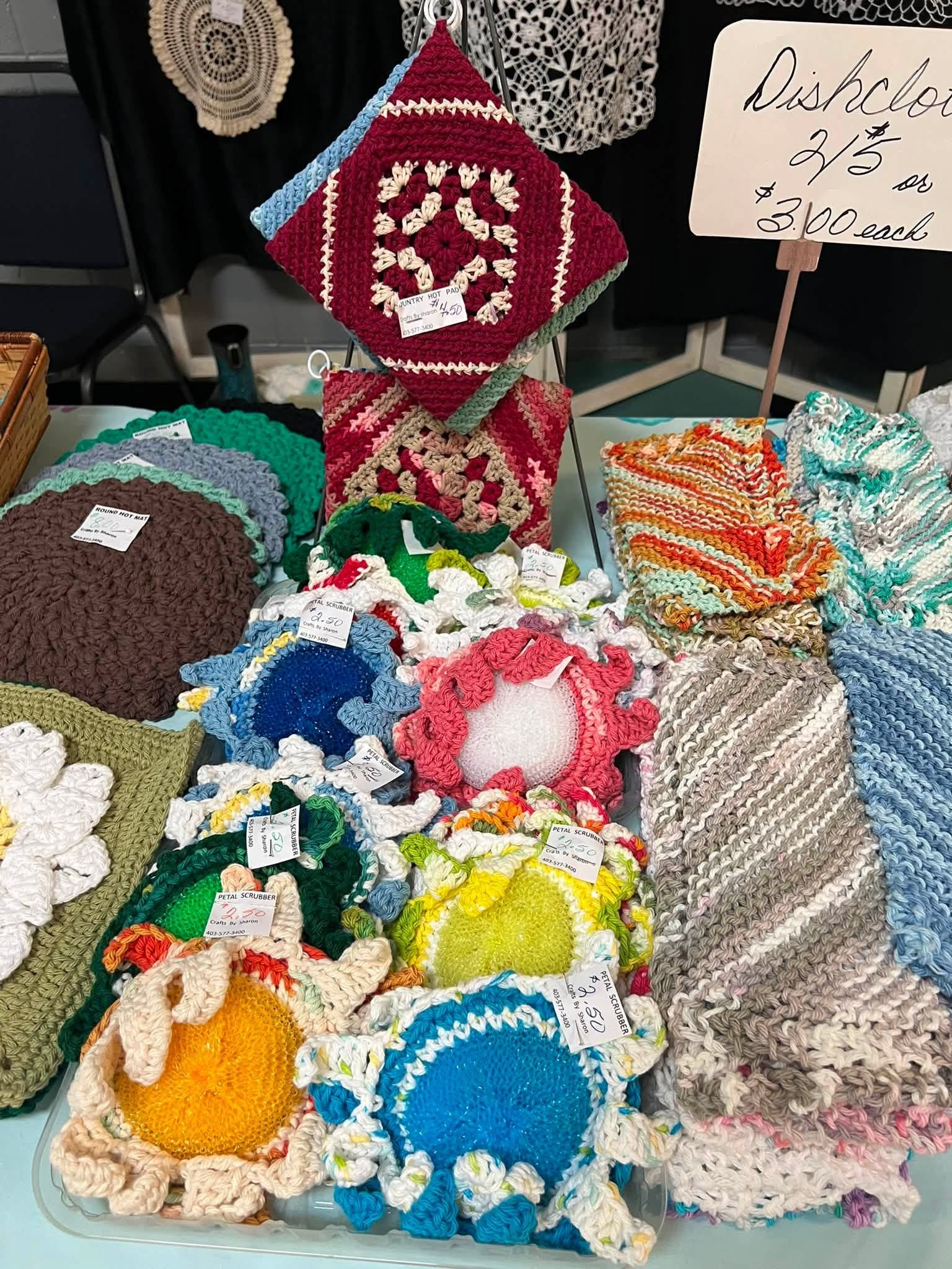 Table display of colourful handmade crocheted dishcloths, pot holders and scrubbers with price tags.