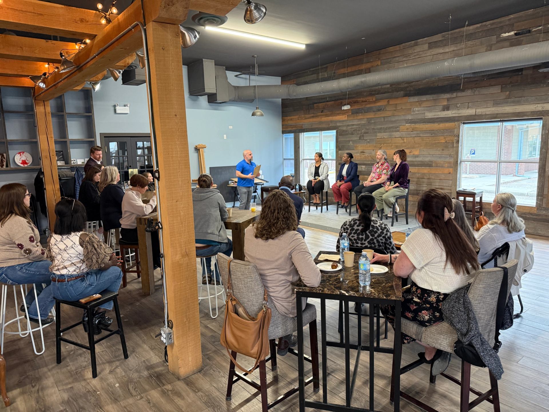 Audience in casual café-style room listens to panel discussion on raised platform.