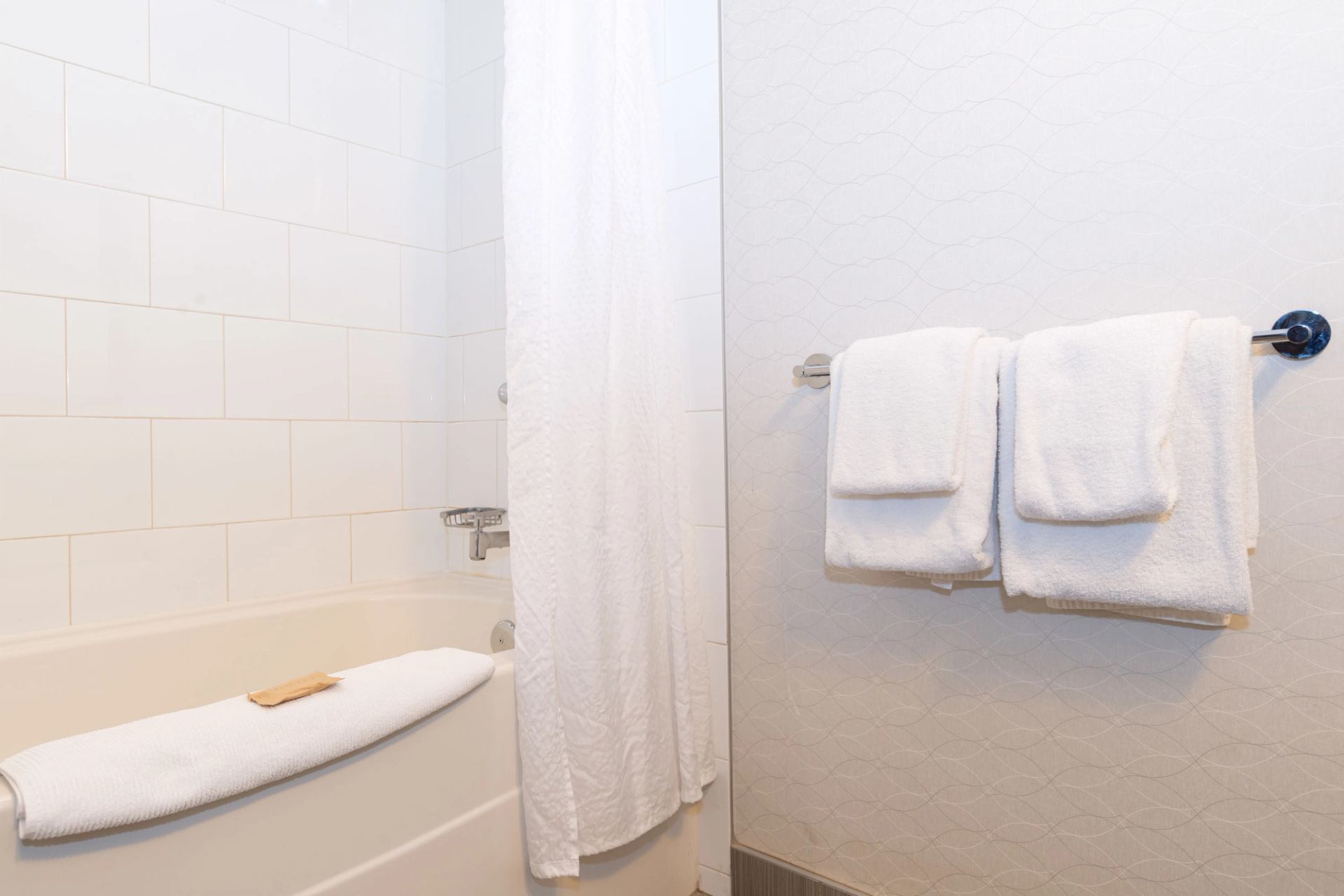 White bathroom with bathtub, shower curtain, folded towels on rack and soap on towel.
