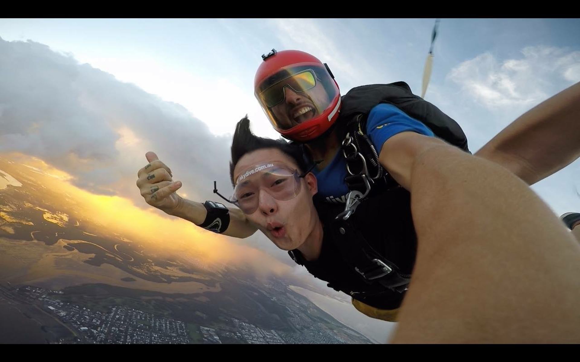 Two people tandem skydiving at sunset with land and water below.