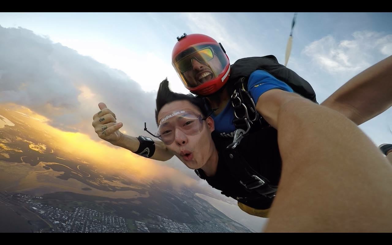Two people tandem skydiving at sunset with land and water below.
