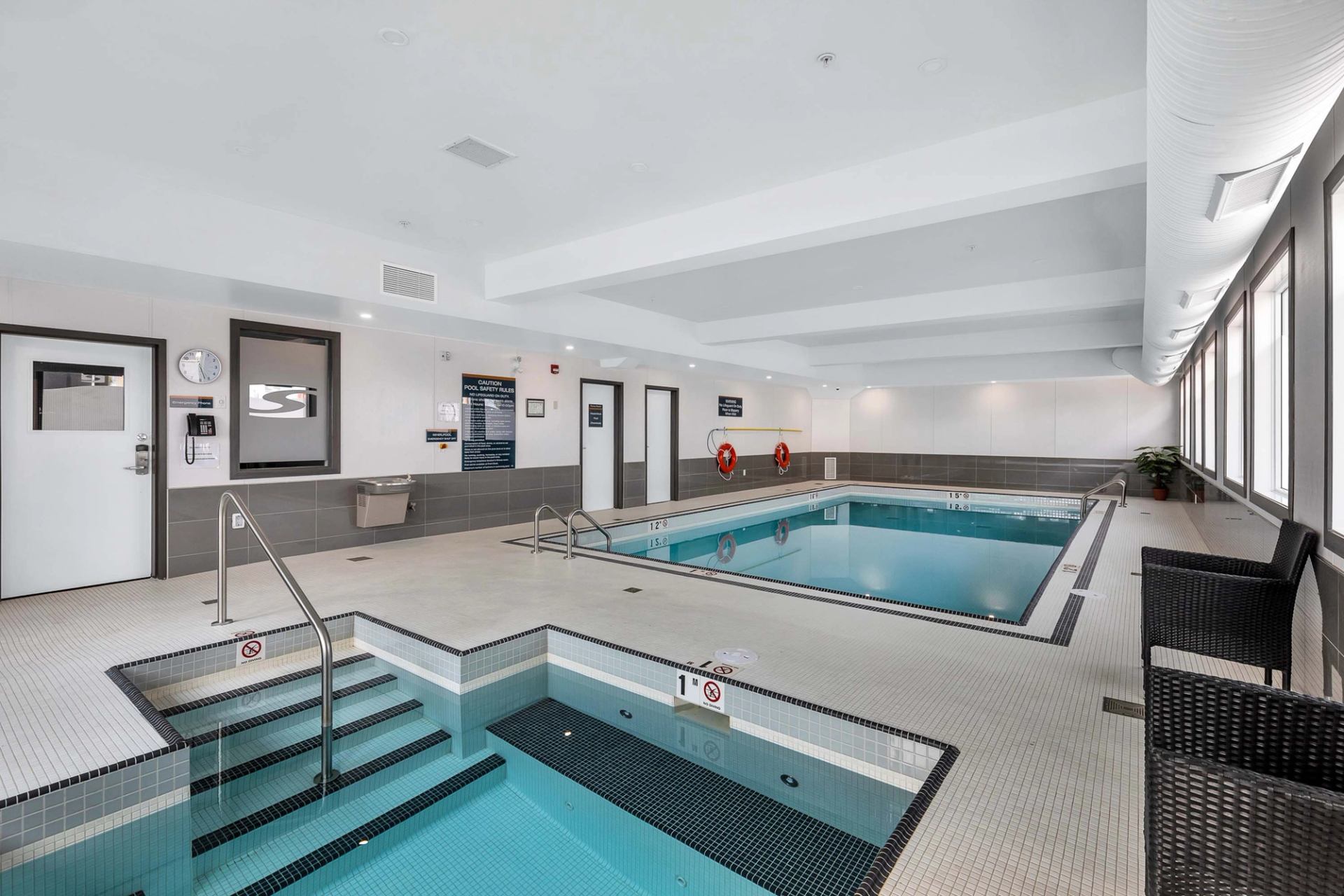 Indoor hotel pool area with hot tub, safety signs, windows and seating.