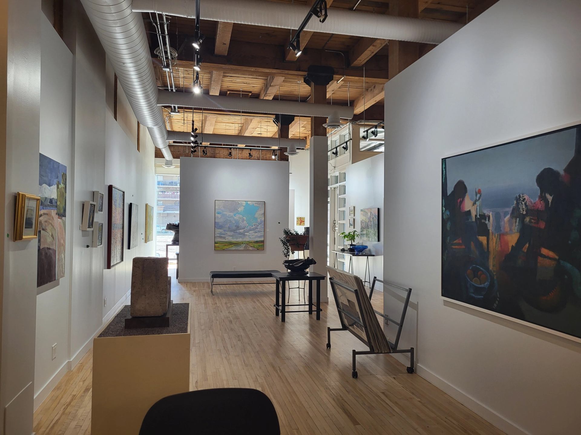 Art gallery interior with paintings, sculptures, wood floors and exposed ceiling ducts.
