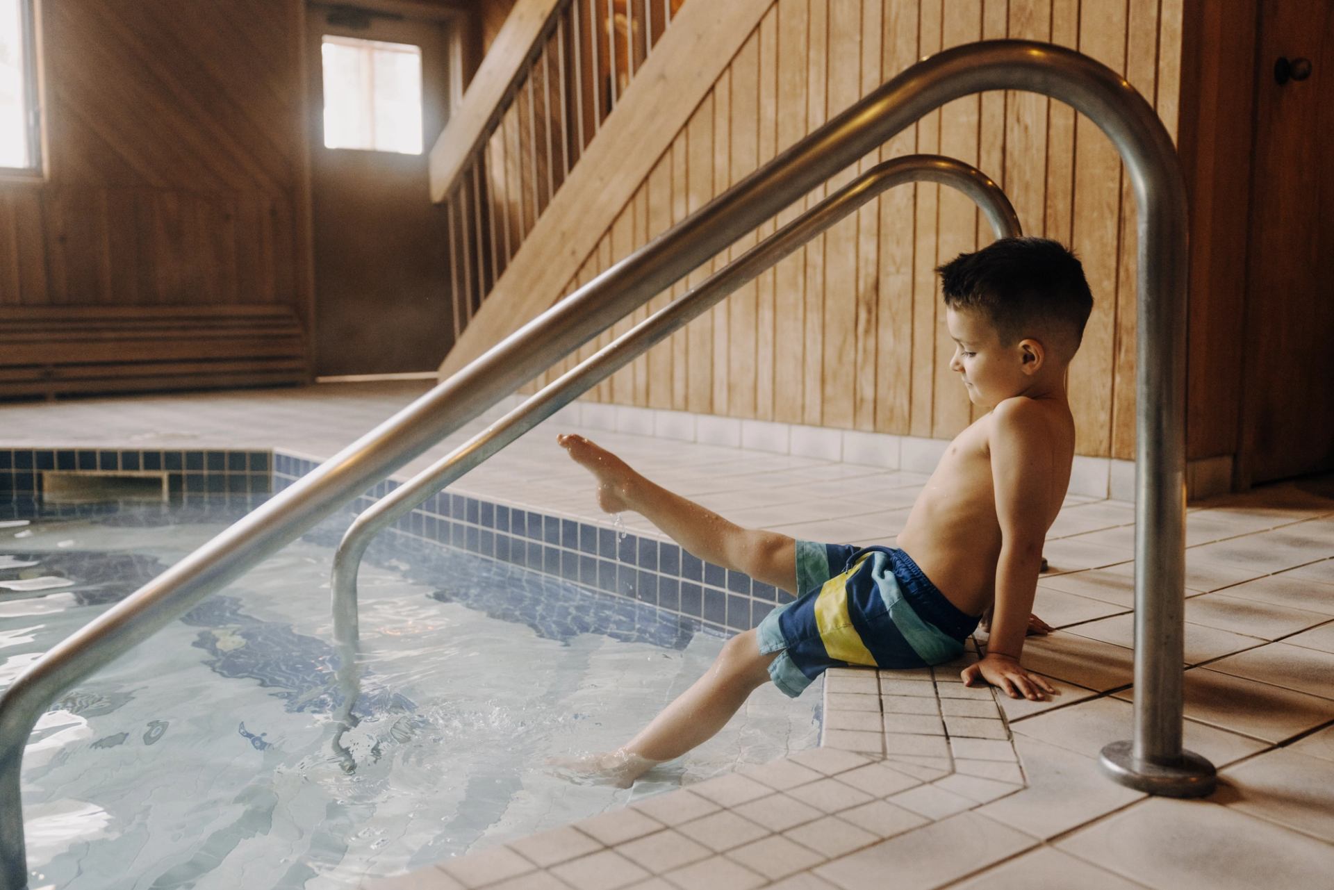 Child sitting at the edge of an indoor pool with feet in the water.