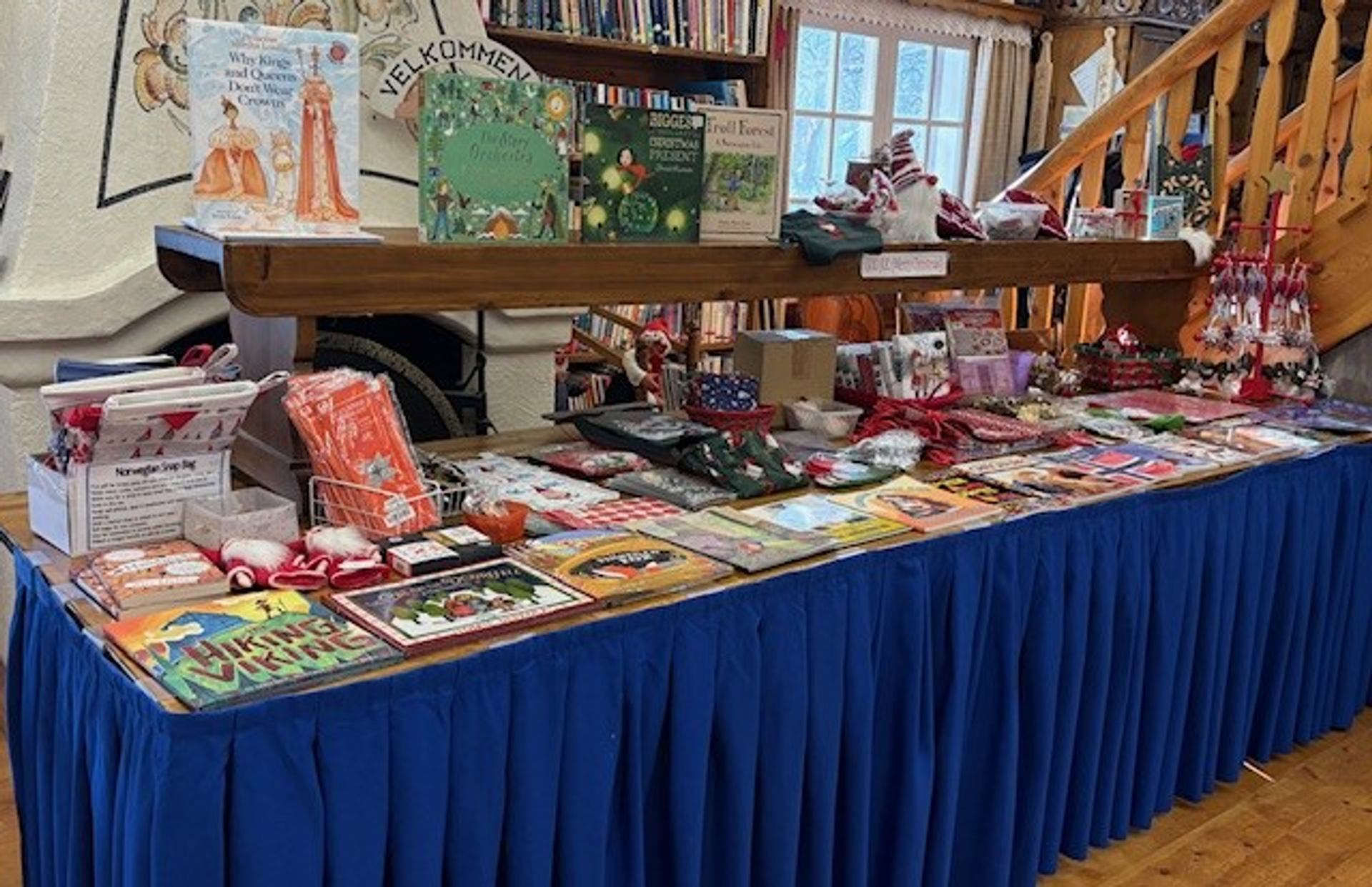 A long blue-draped table laden with books and holiday-themed merchandise in a room with bookshelves and a wooden staircase.