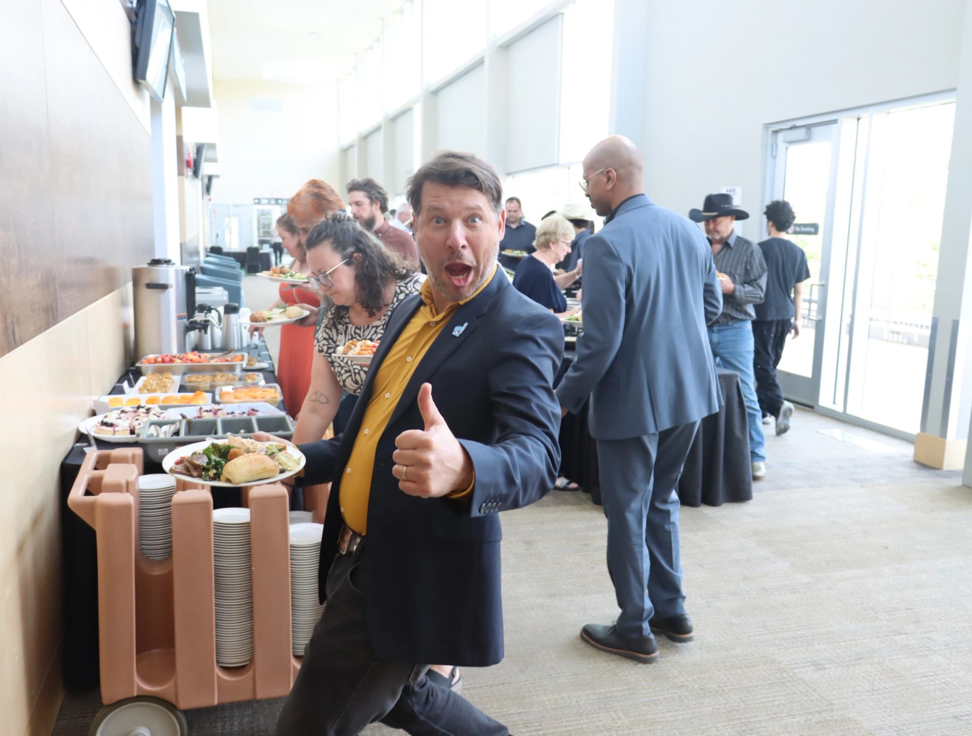 Man holding a full plate gives thumbs-up near buffet table at indoor event.