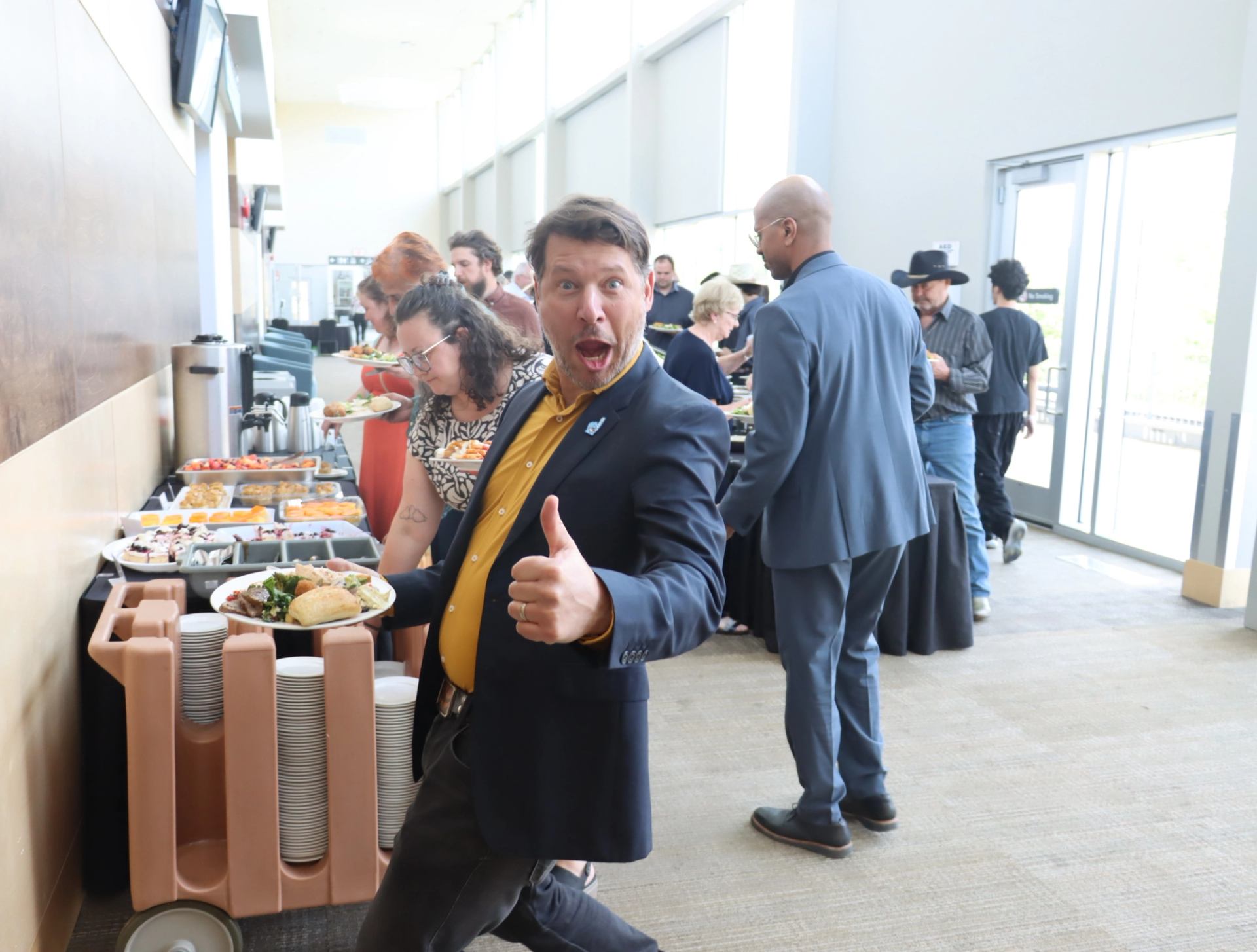 Man holding a full plate gives thumbs-up near buffet table at indoor event.