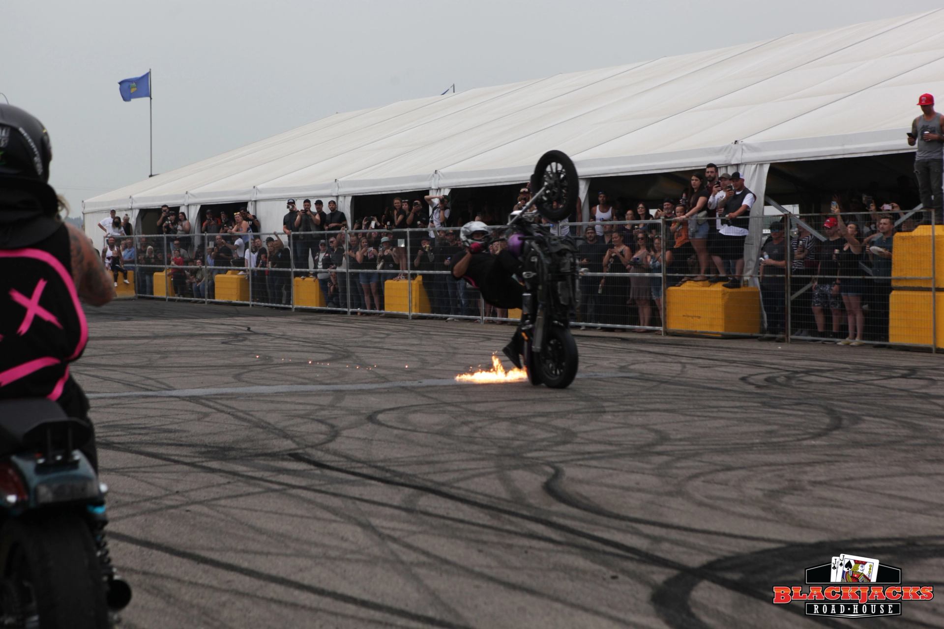Motorcycle stunt show performer rides black motorcycle with flames near track, spectators behind fen.