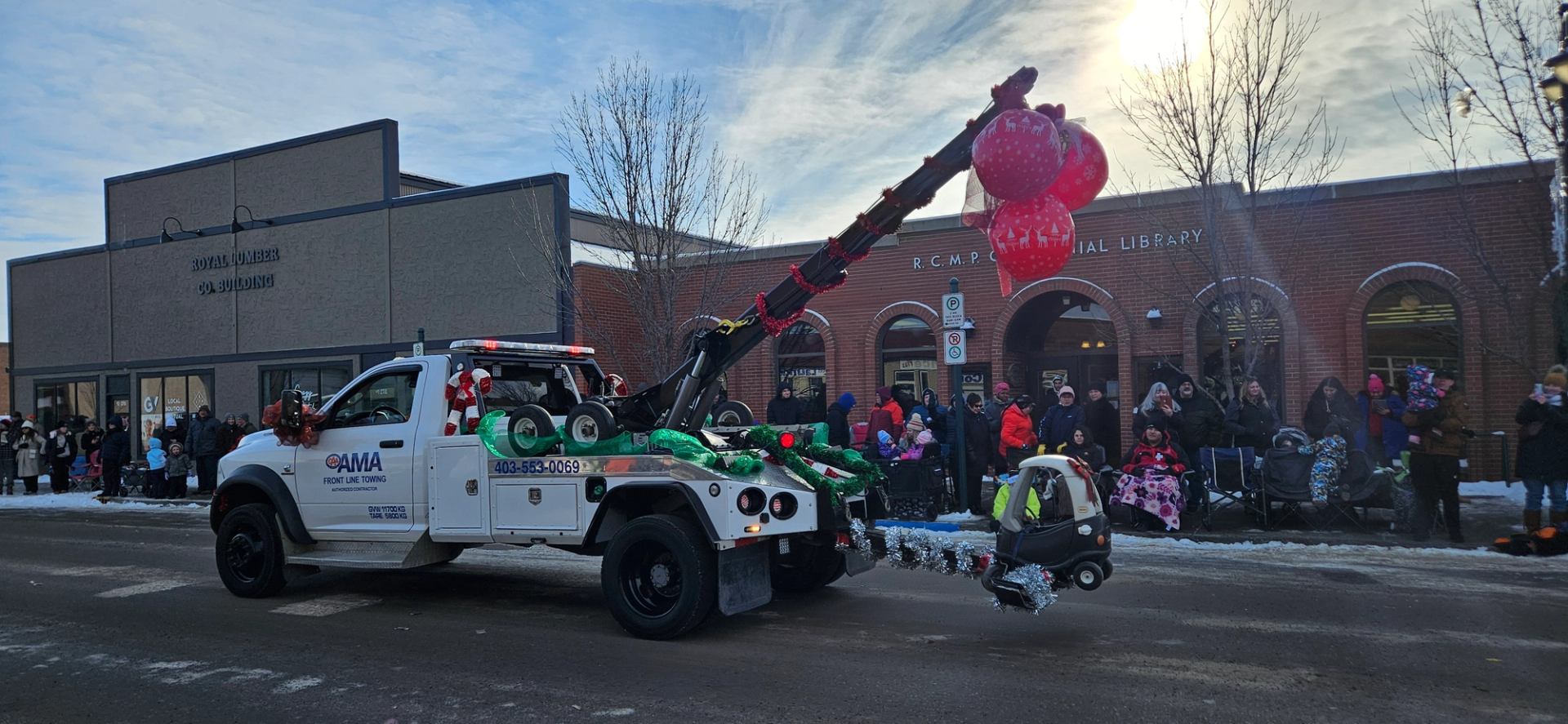 Festive tow truck with balloons and garlands drives in snowy parade past cheering crowd.