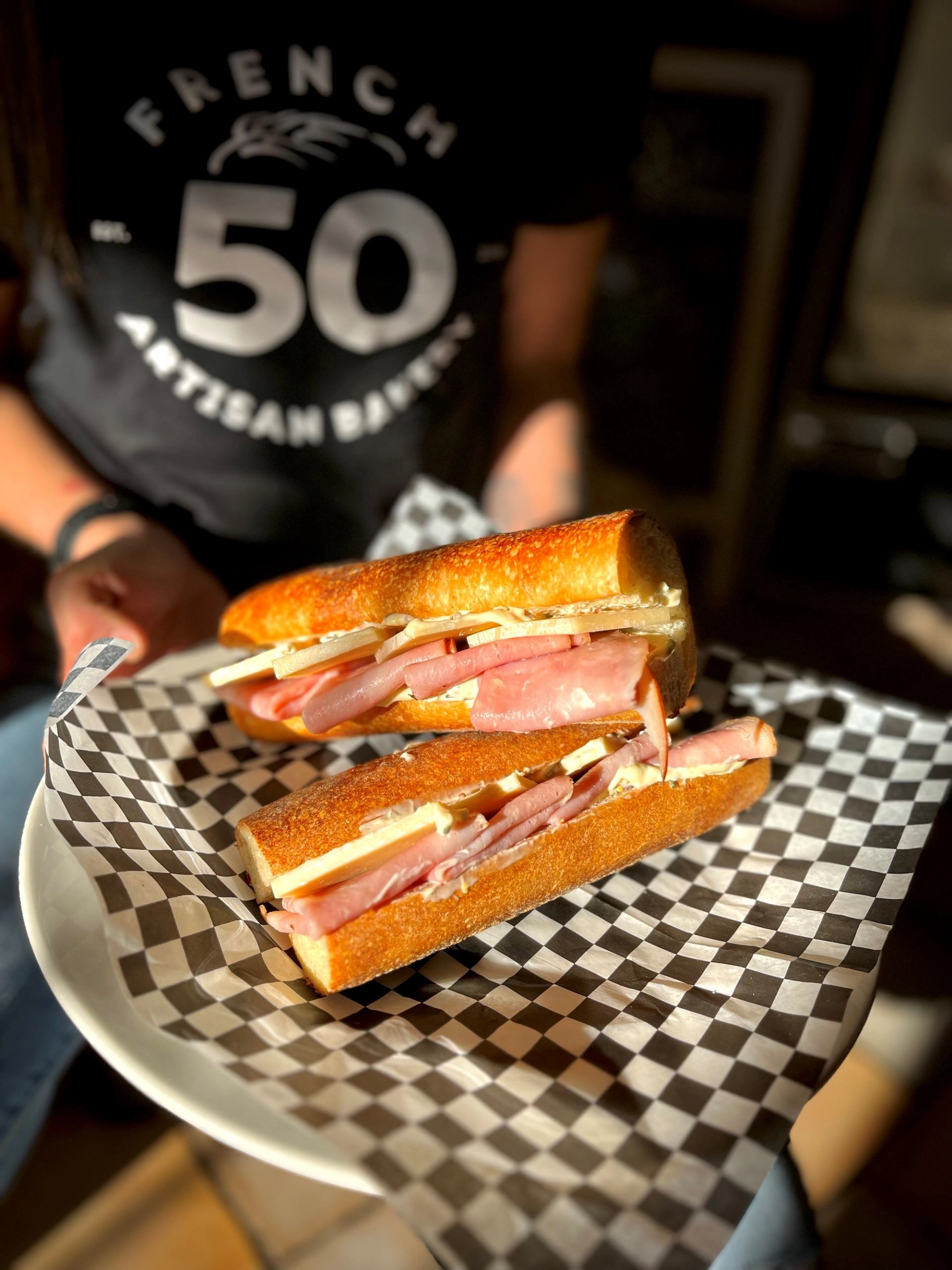 Sandwiches with ham and cheese on a checkered tray, French 50 Bakery sign in background.