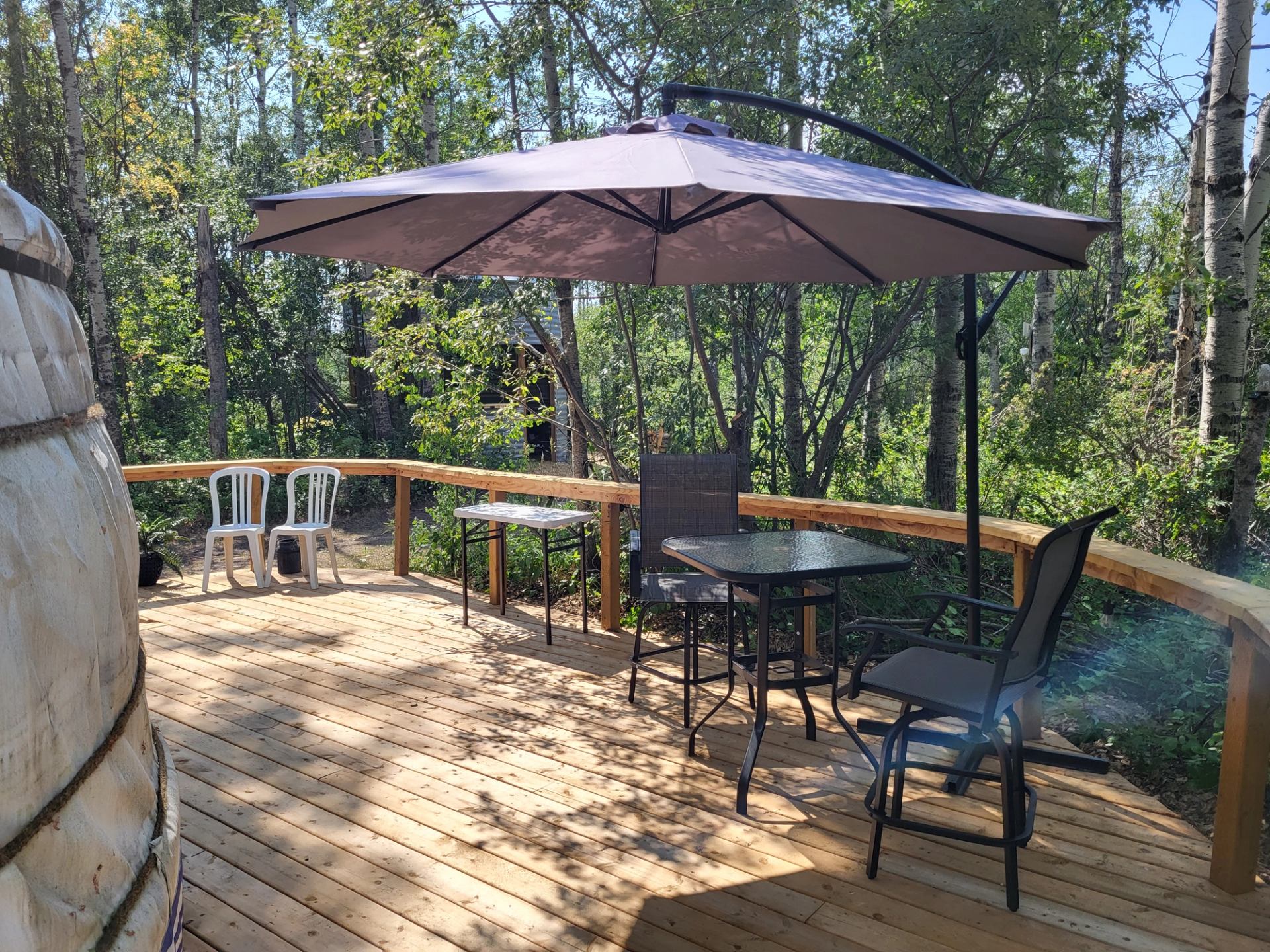A wooden deck with patio furniture and a large umbrella, surrounded by trees, with a yurt partially visible on the left.