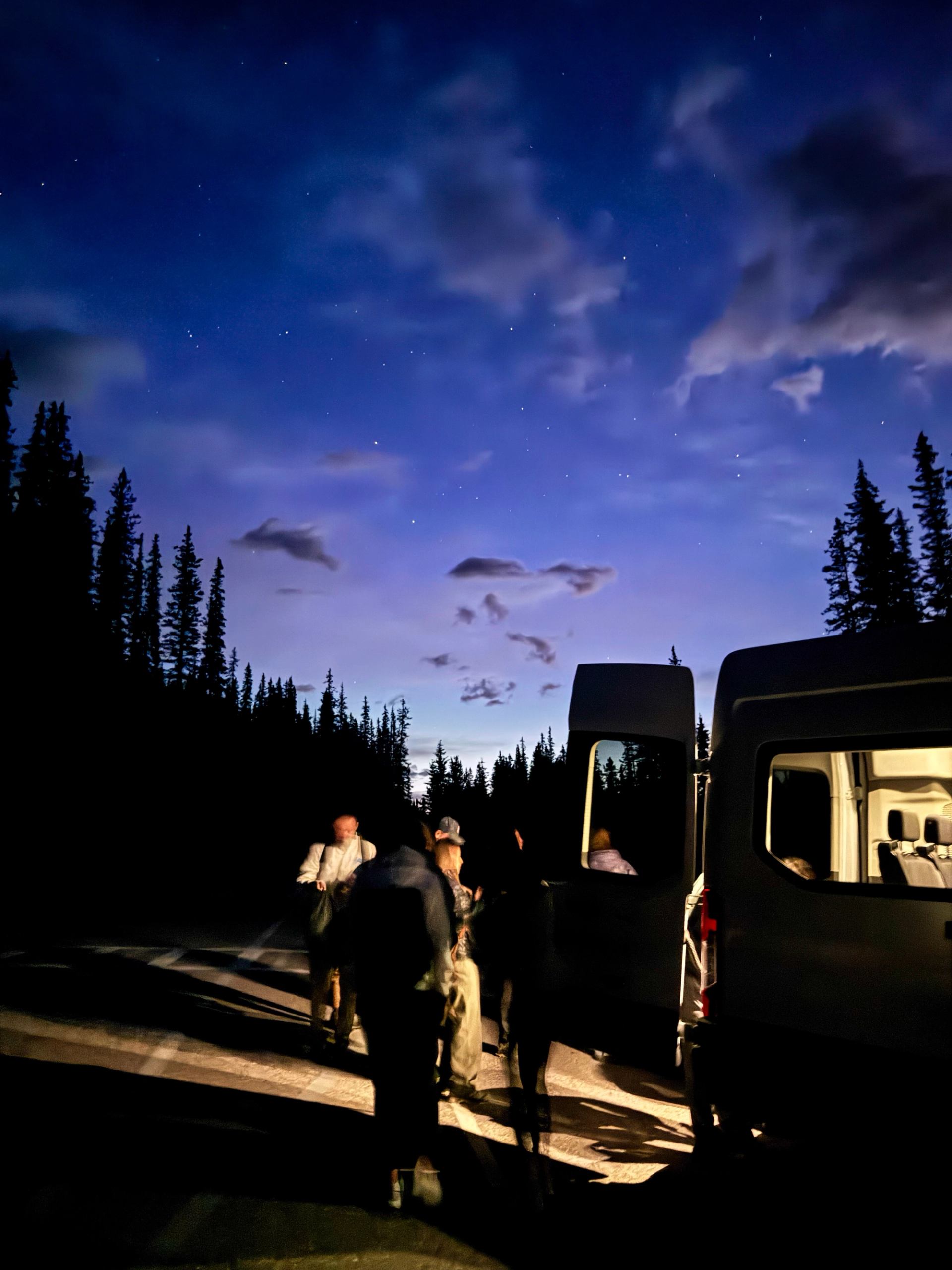 People standing near a van under a twilight sky with trees silhouetted around them.