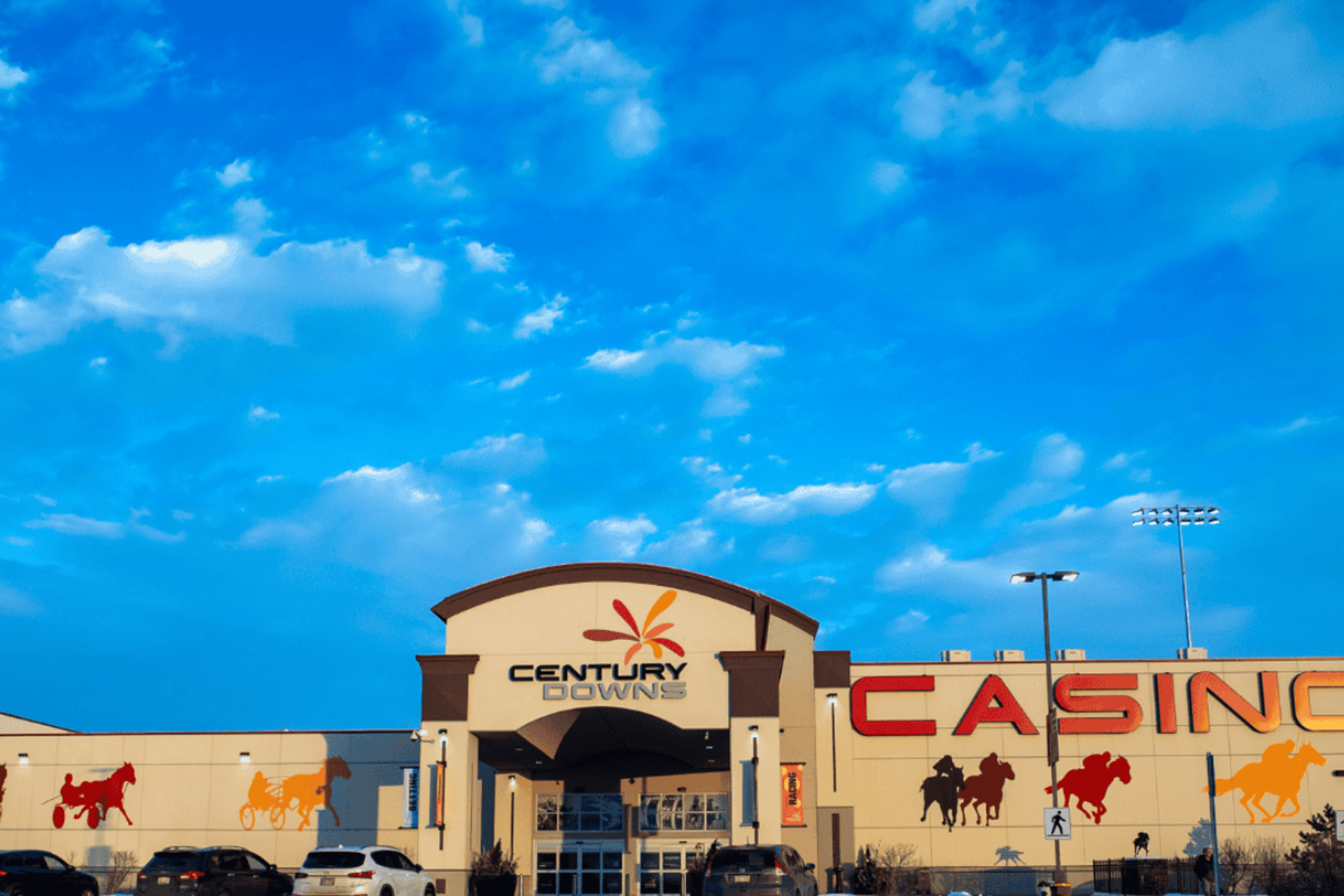 Century Downs casino exterior with horse racing murals and parked cars under blue sky.
